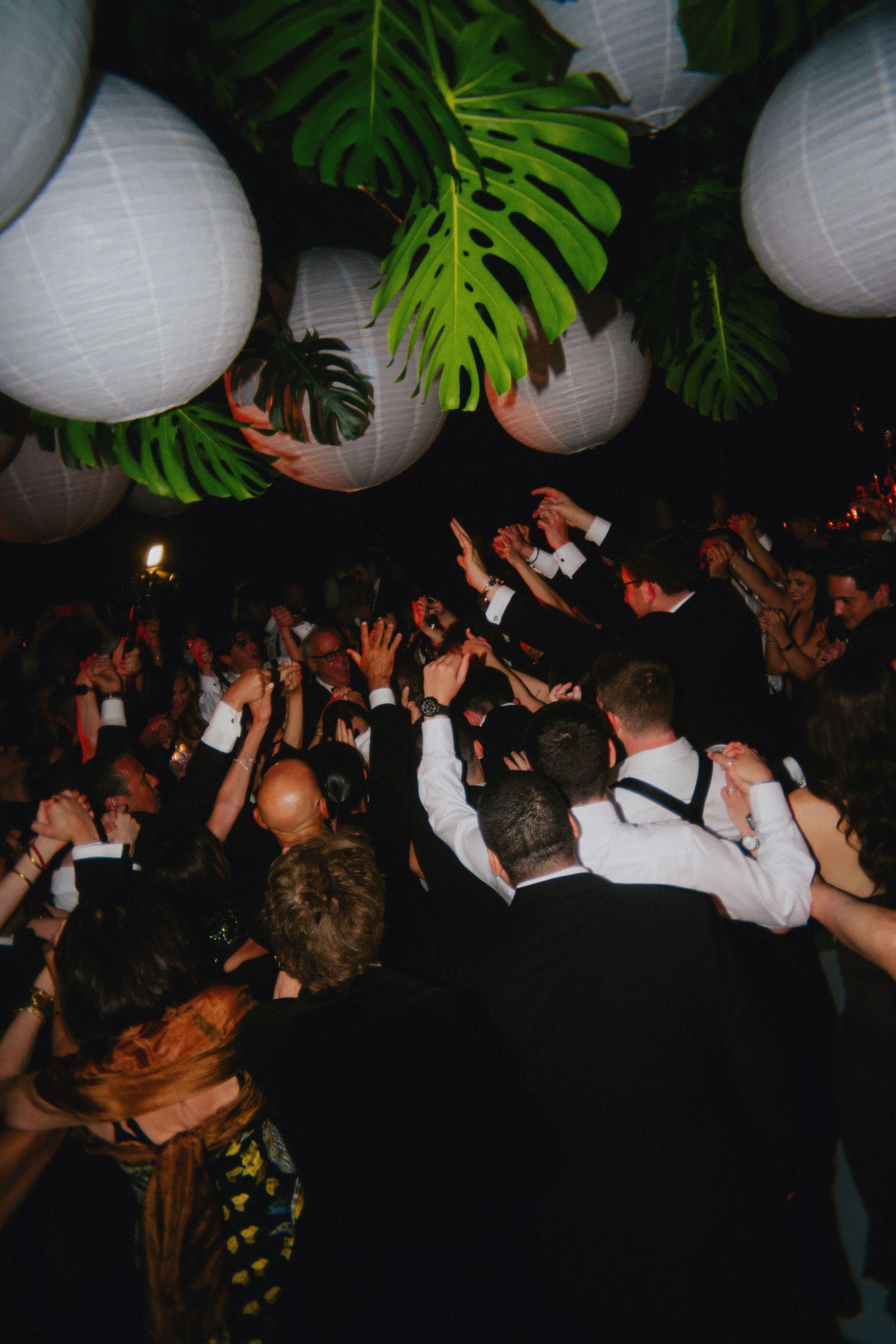 Lively wedding reception dance floor with guests celebrating under white paper lanterns and tropical monstera leaf decorations.