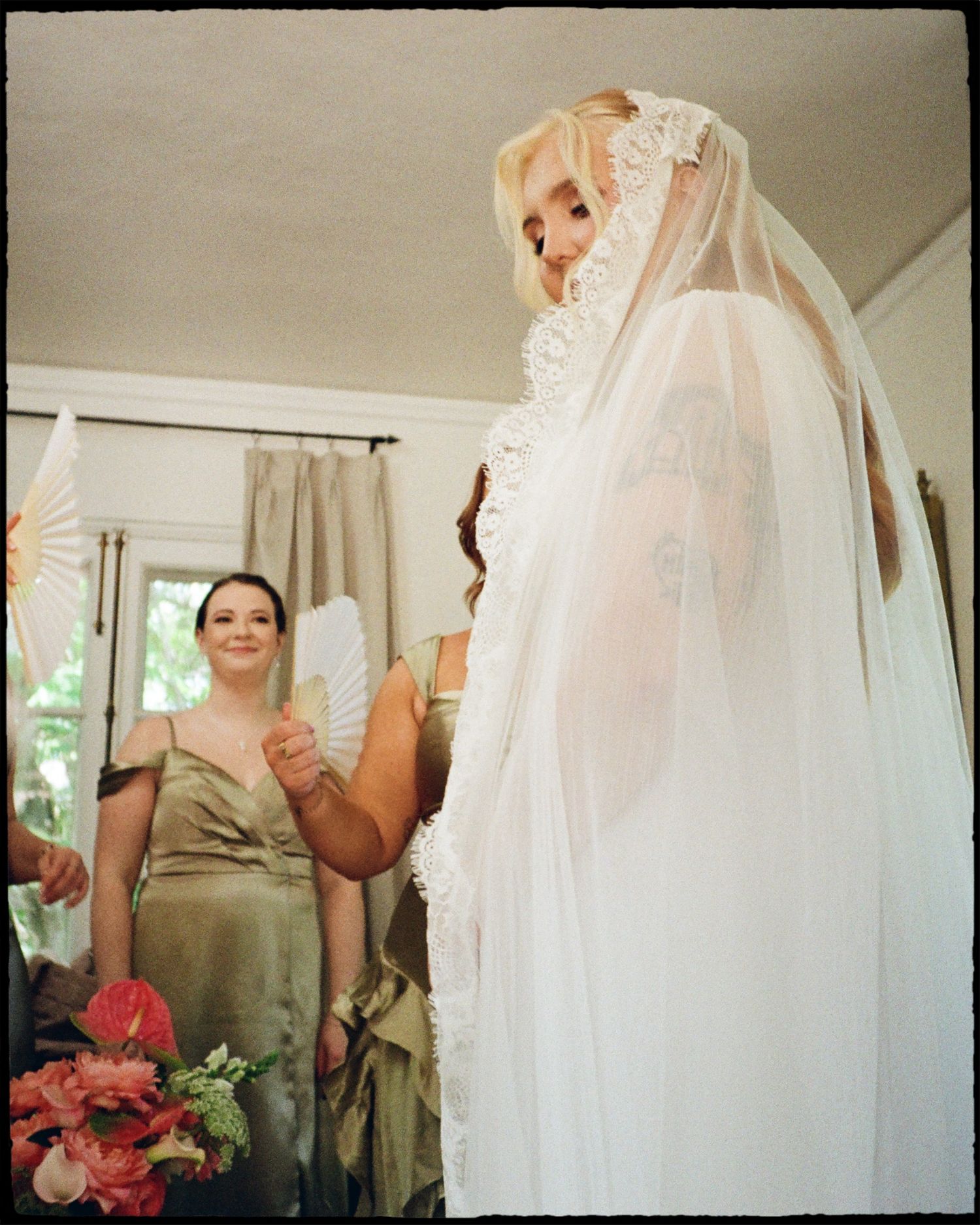 A bride in a white lace veil holds hands with a bridesmaid in a green dress holding red flowers indoors.