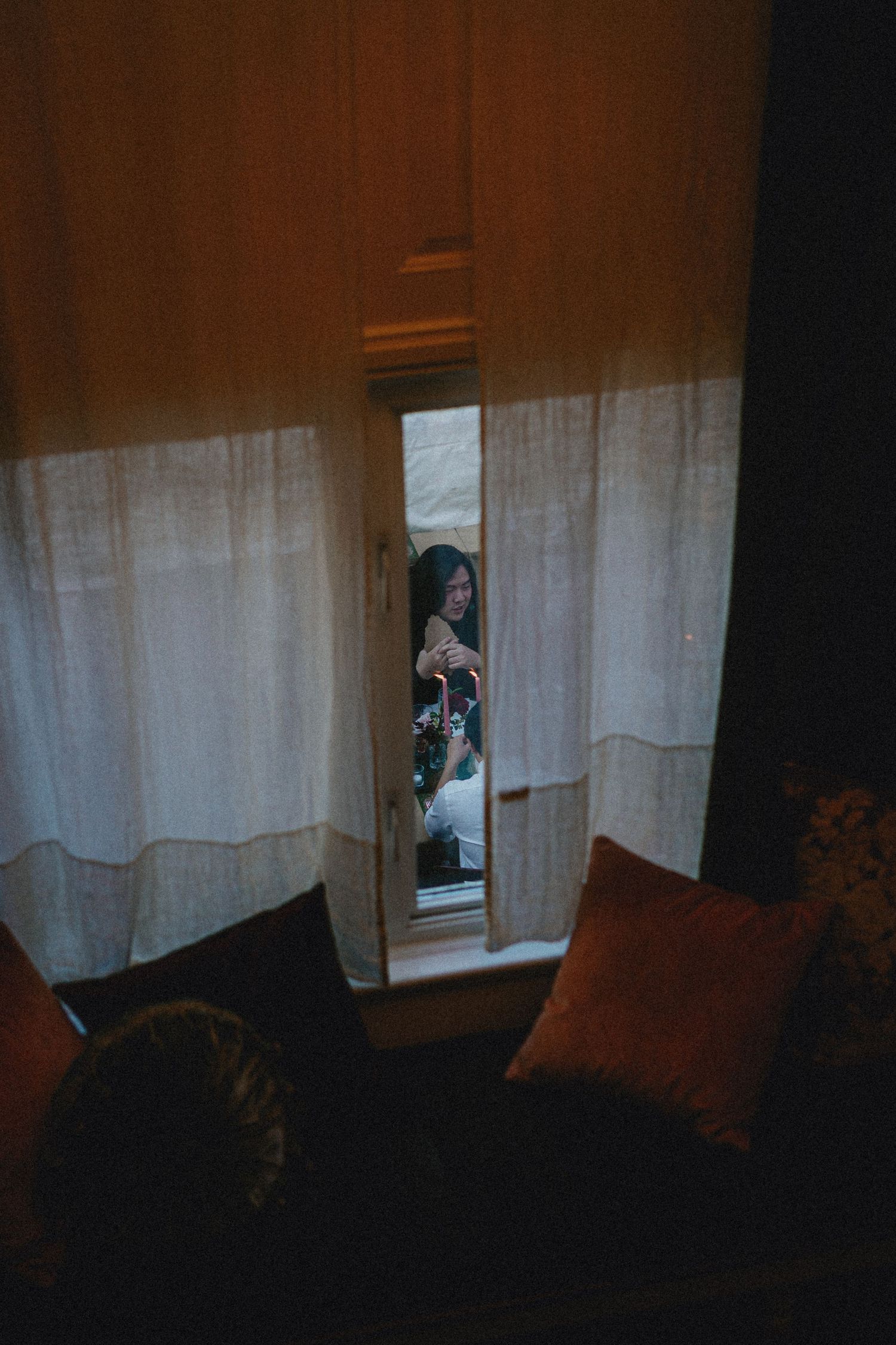 Woman reflected in a narrow mirror between sheer white curtains in a dimly lit room with orange velvet pillows.