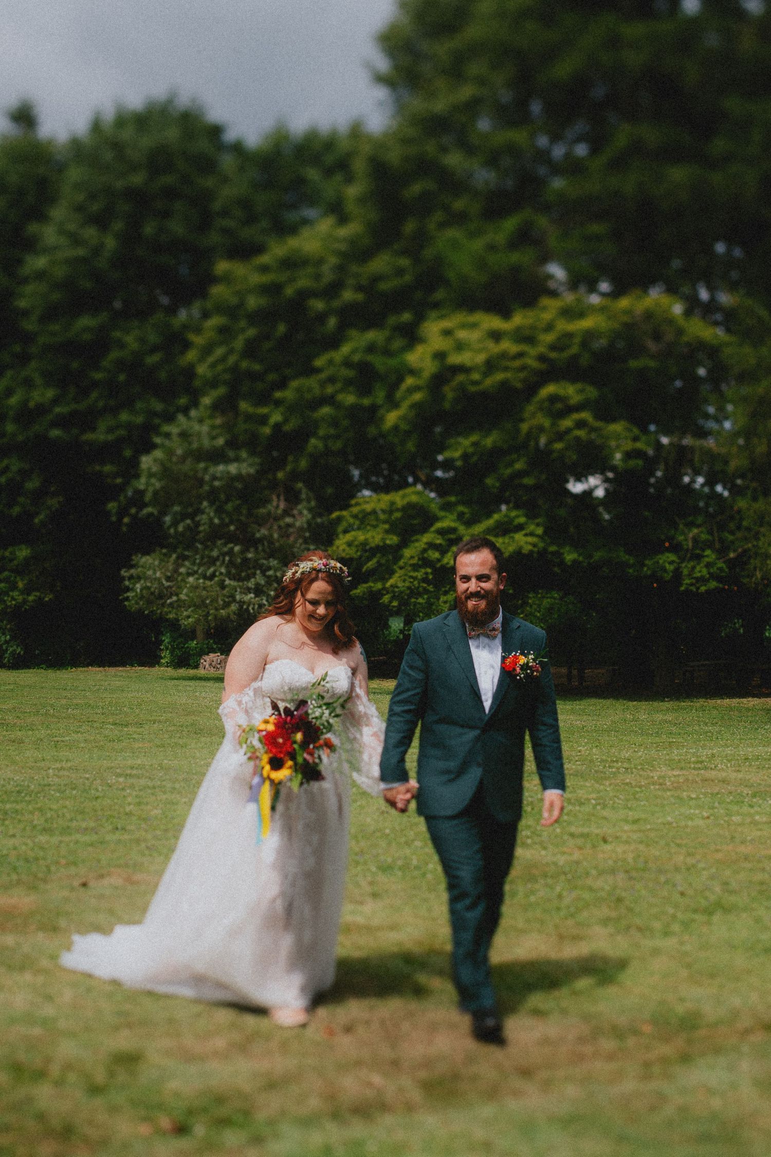 Bride and groom walk hand-in-hand across lush green lawn, she in white gown with colorful bouquet, he in teal suit.