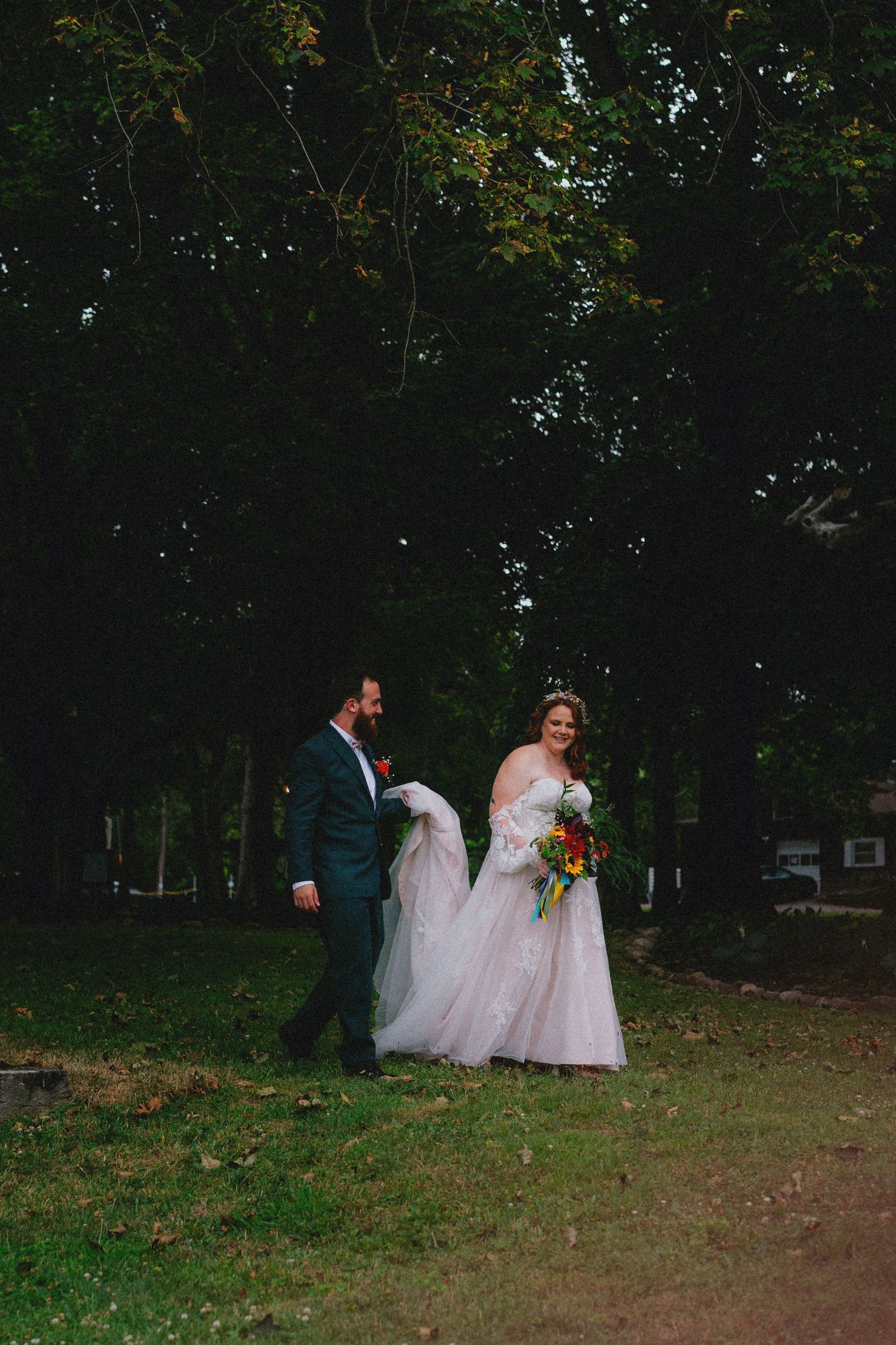 Bride and groom walk hand-in-hand outdoors, she holds colorful bouquet in white gown, he wears navy suit among lush green trees.