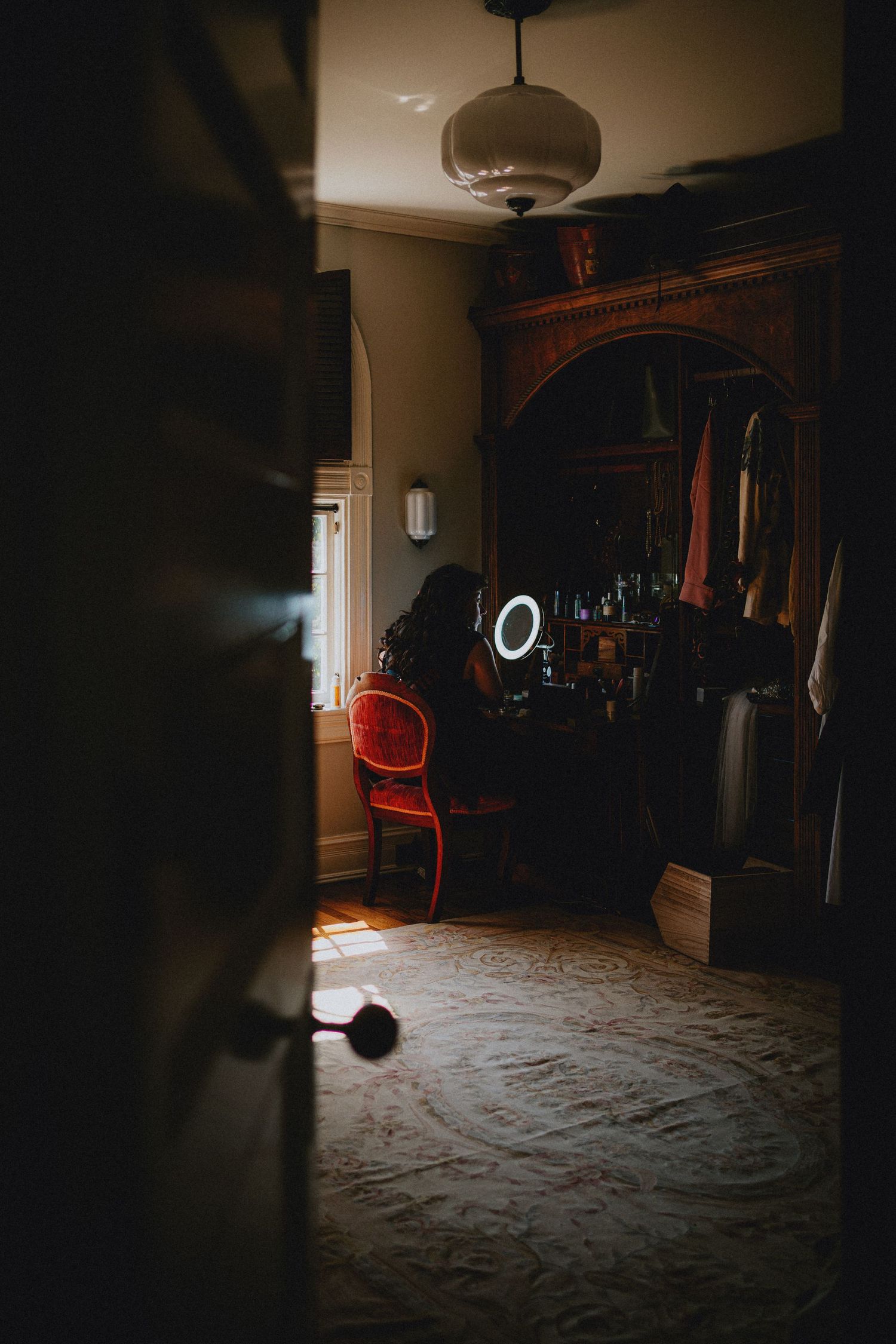 Person sitting at a vanity mirror with ring light in a dimly lit vintage bedroom with ornate wooden furniture.