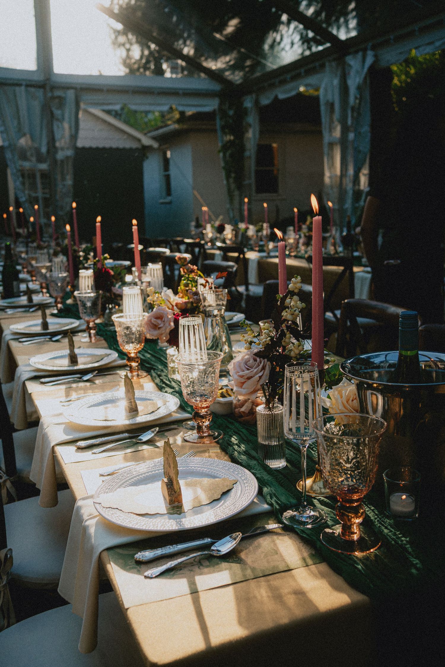 Elegant outdoor wedding reception table with pink taper candles, floral centerpieces, gold accents, and green velvet runner at dusk.