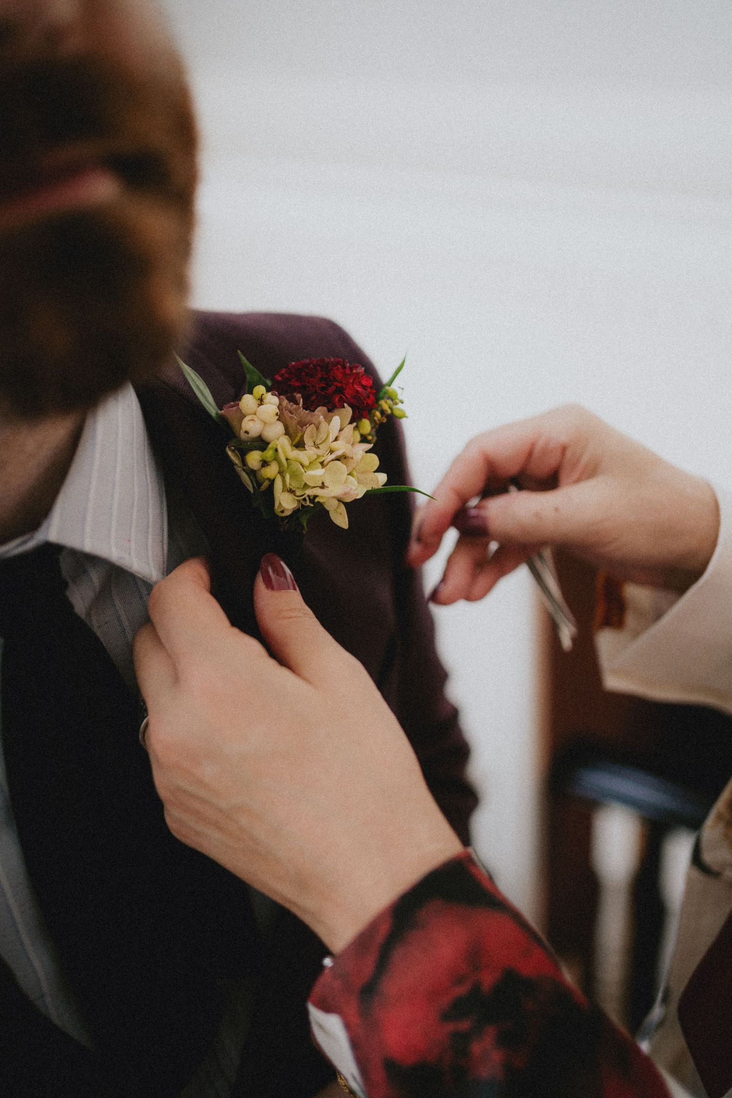 Hands pinning a floral boutonniere onto a groom's burgundy suit lapel at a wedding.