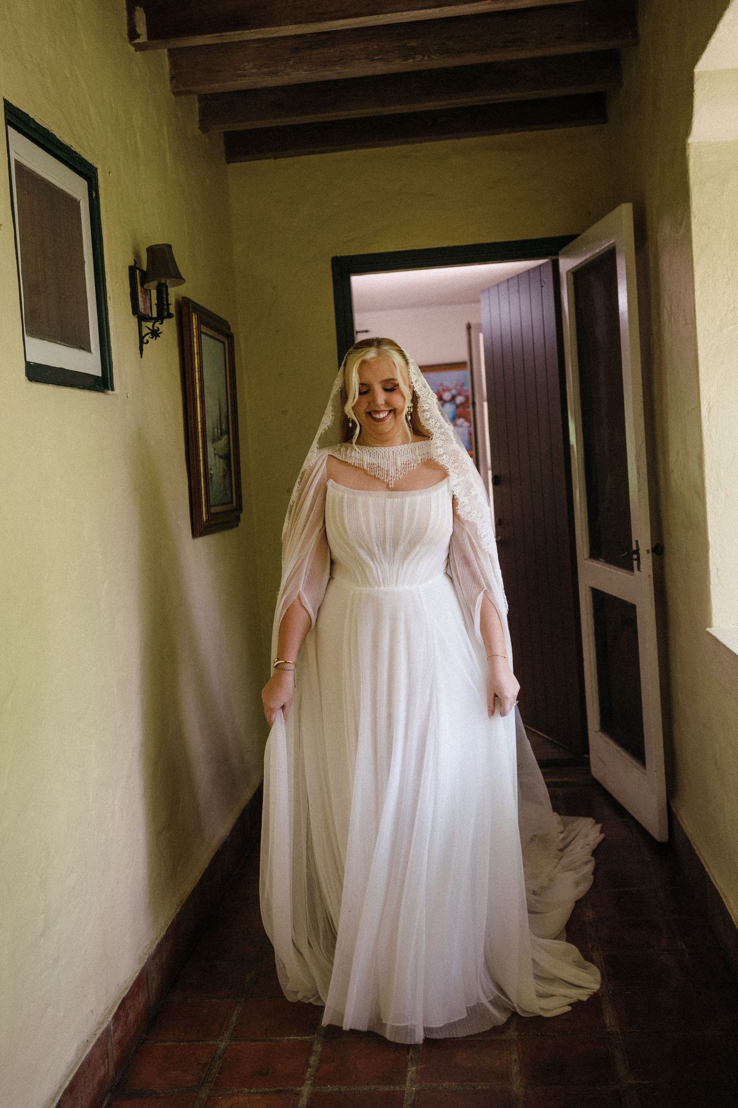 Smiling bride in a flowing white wedding gown with veil stands in a rustic hallway with green walls and wooden beams.