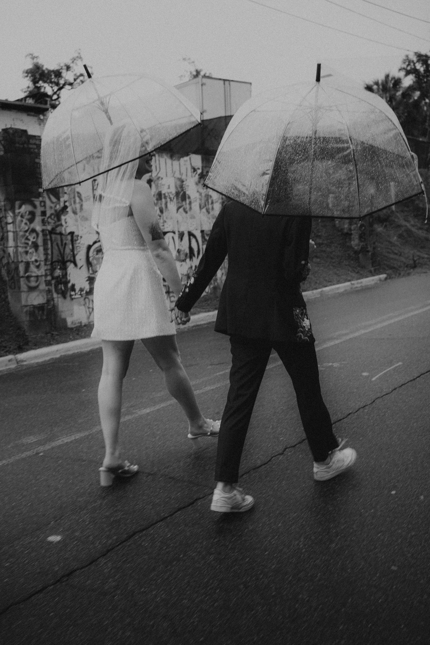 Black and white photo of a couple holding hands and walking in the rain under clear bubble umbrellas, dressed in wedding attire.
