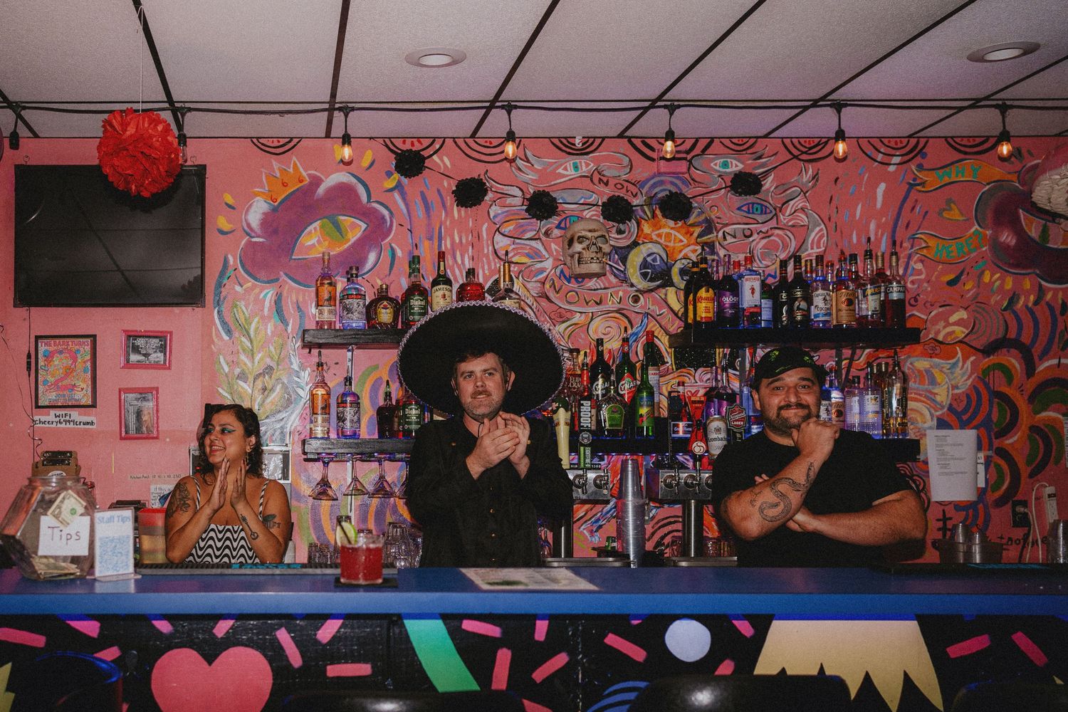 Three bartenders pose behind a vibrant, colorfully painted bar stocked with liquor bottles in a lively venue.