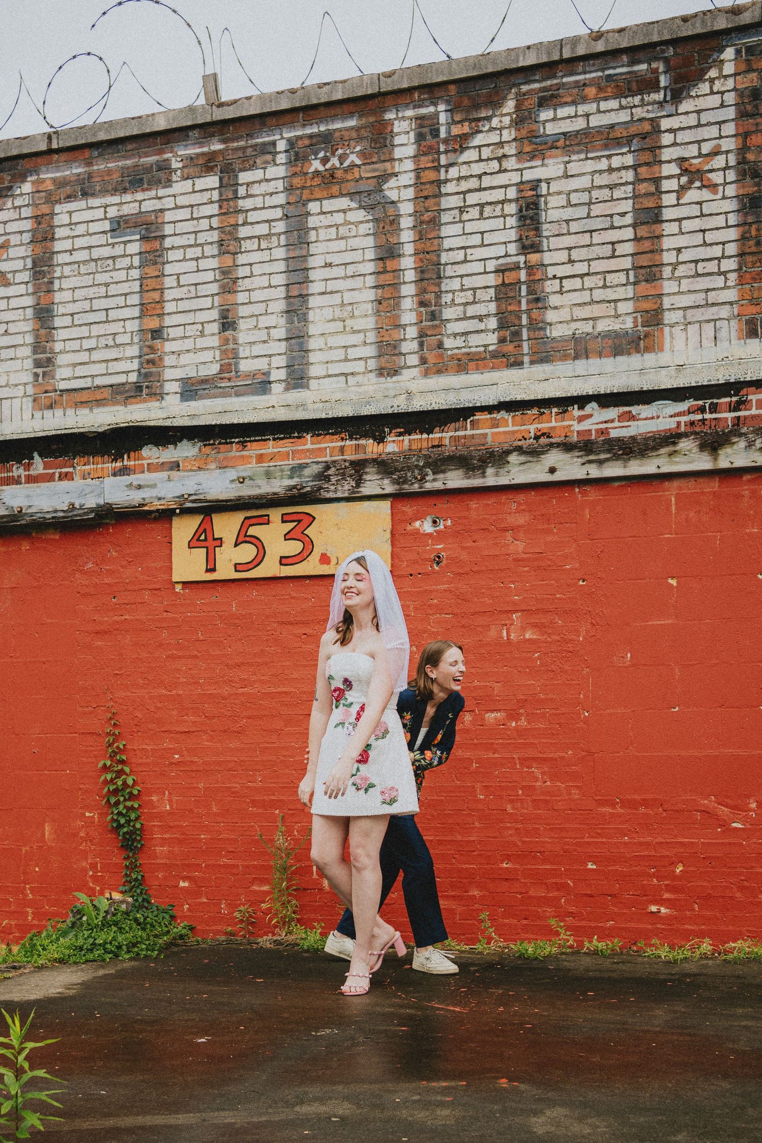 Bride in floral dress and veil poses with friend against red brick wall with number 453 painted on it.