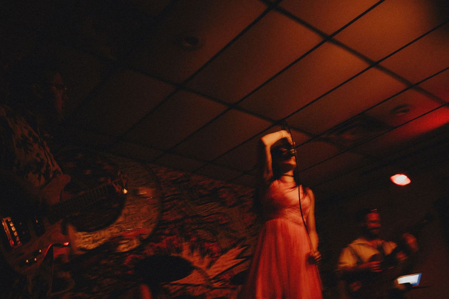 Woman in pink dress performing on stage with microphone under red ambient lighting in a dimly lit venue.