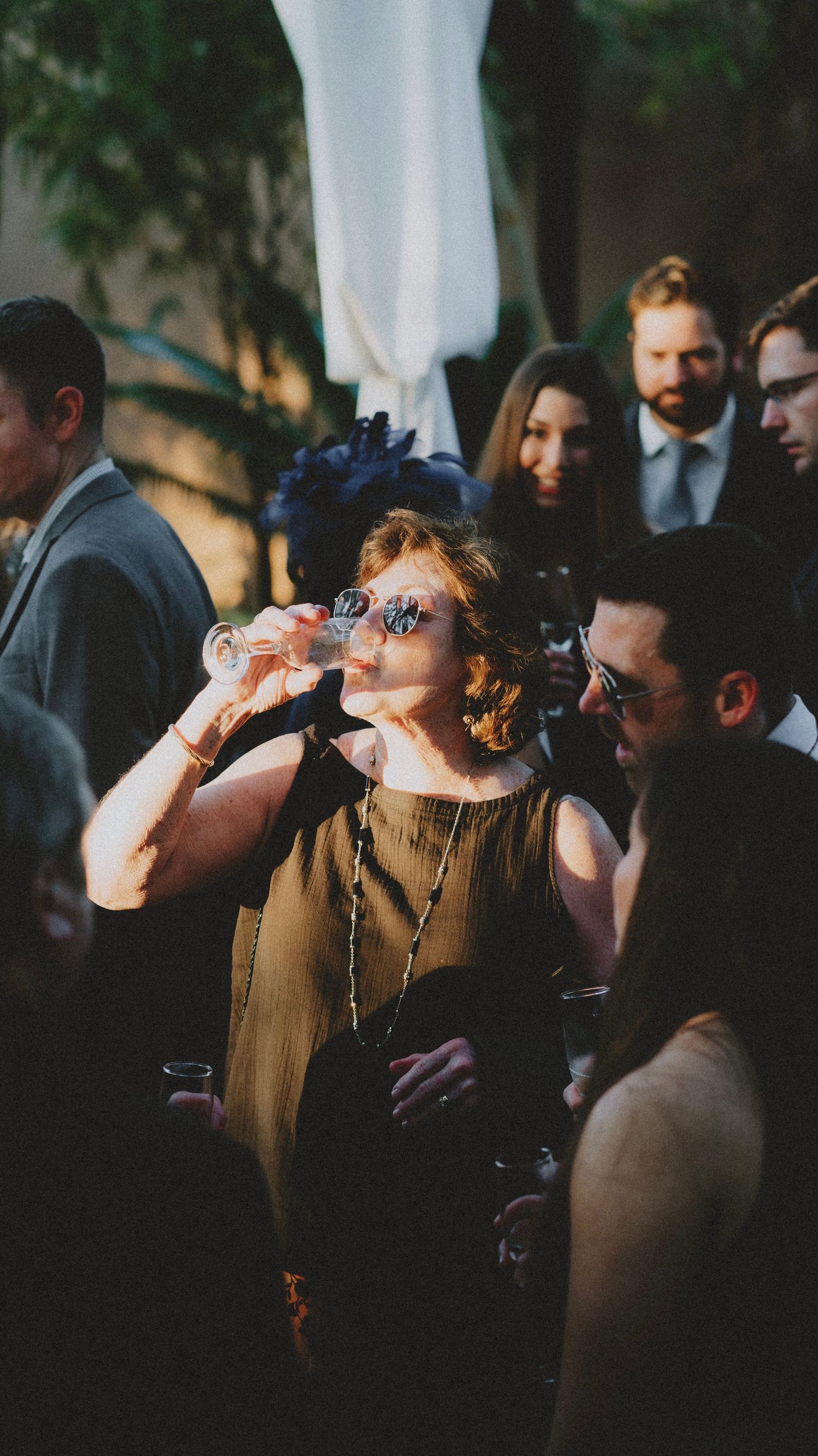 Elegantly dressed woman drinking champagne at an outdoor evening party surrounded by well-dressed guests.