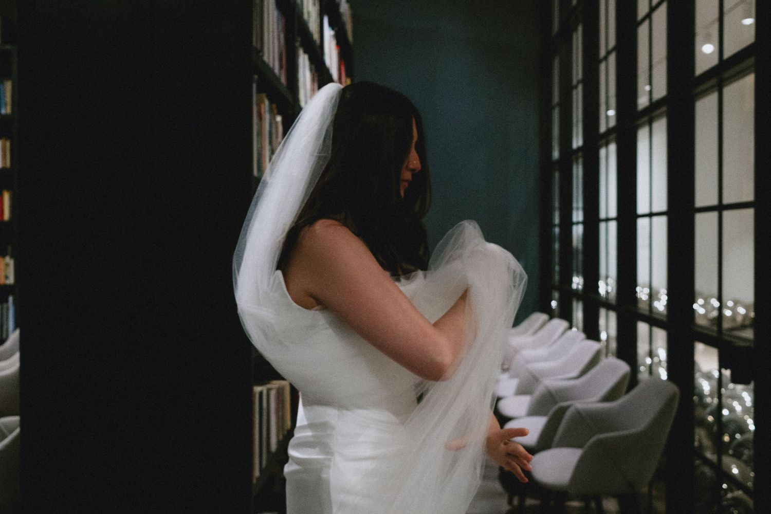 Bride in white wedding dress and veil standing elegantly in a dimly lit room with large windows.