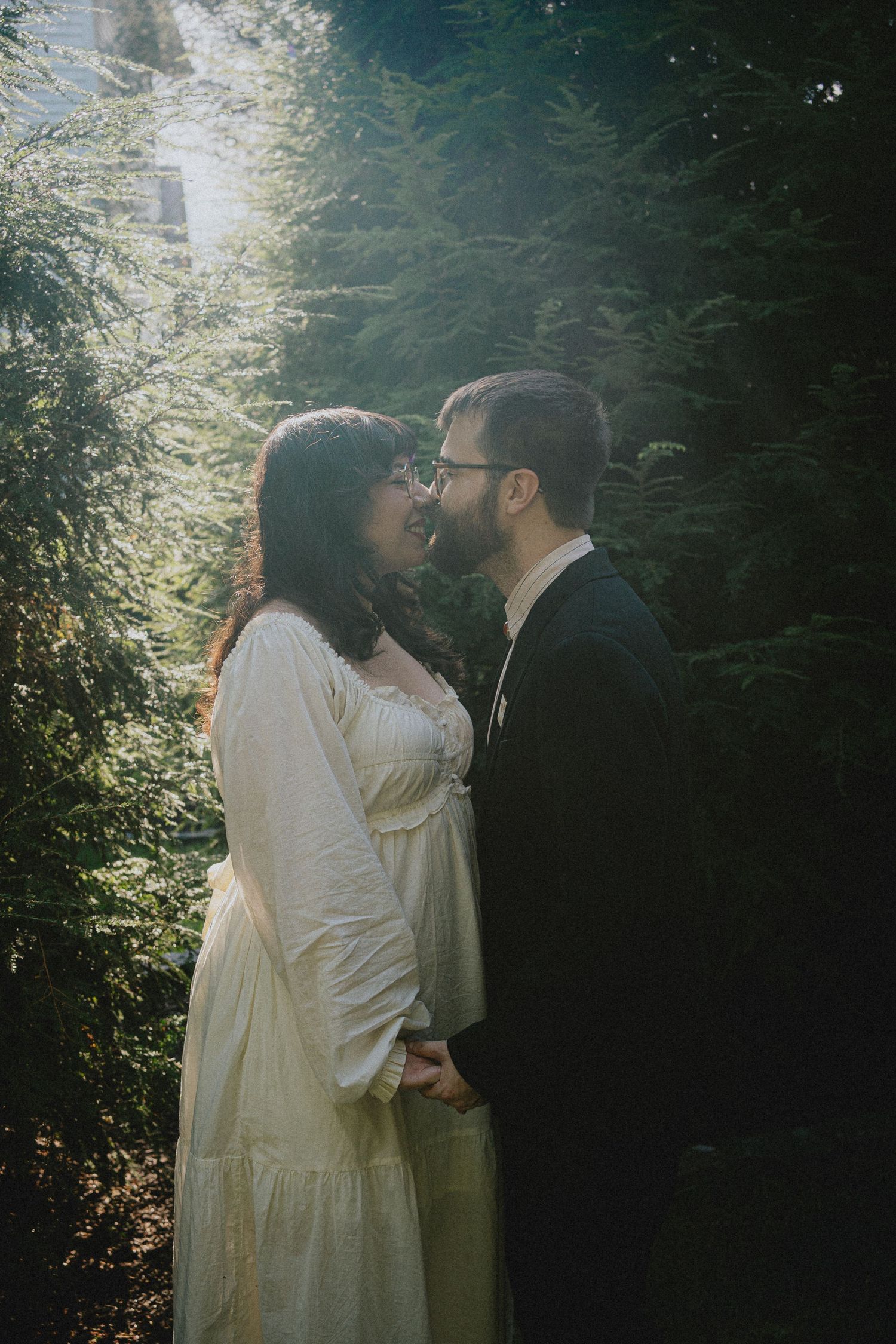 Couple sharing a romantic moment in a sunlit forest, bride in white dress and groom in dark suit holding hands.