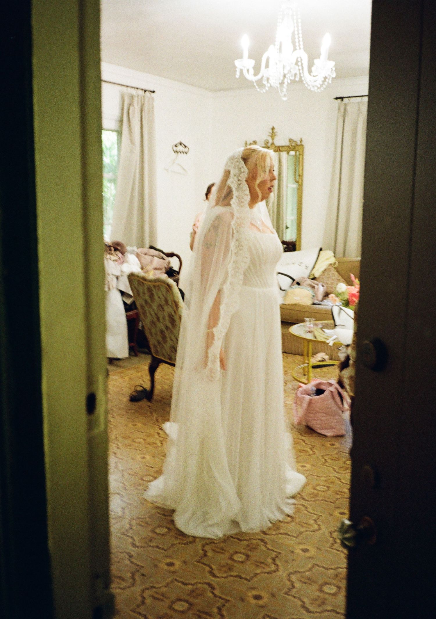 Bride in white wedding gown and veil stands in elegantly decorated room with chandelier overhead.
