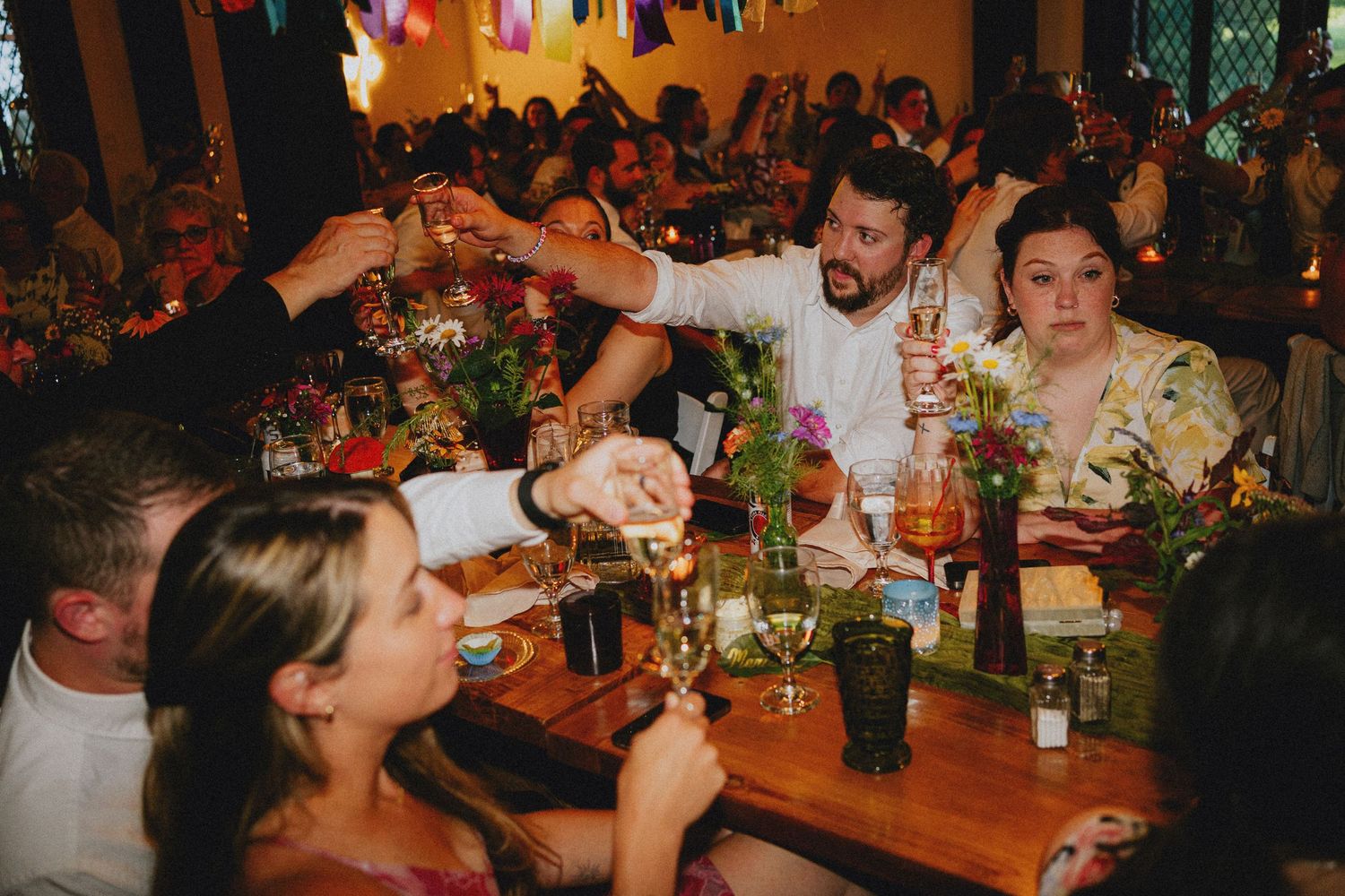 Guests raising glasses in a toast at a colorful wedding reception dinner, seated at long wooden tables with floral centerpieces.