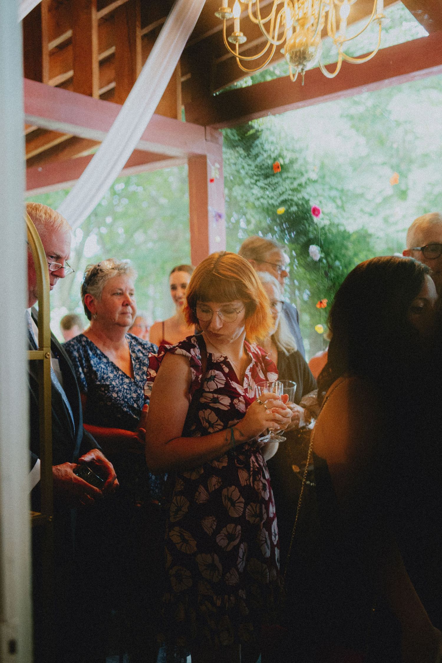 Guests mingle at an outdoor wedding reception under a wooden pavilion with chandelier lighting and lush greenery.
