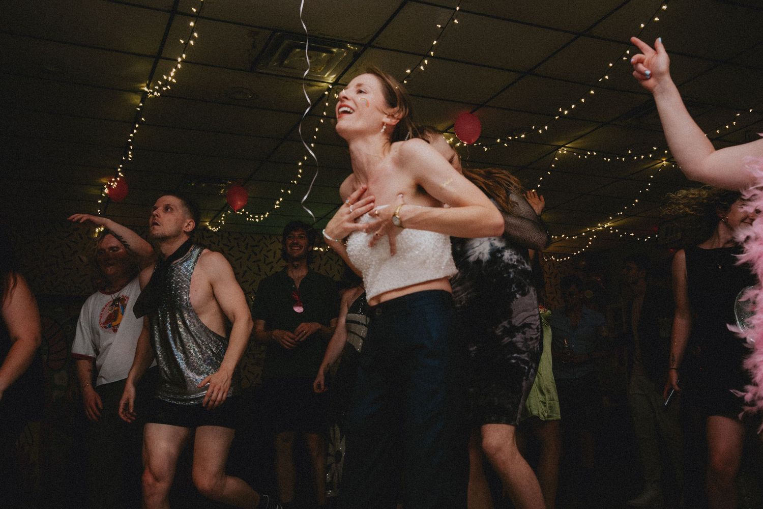 People dancing energetically at an indoor party with string lights decorating the ceiling in a festive venue.