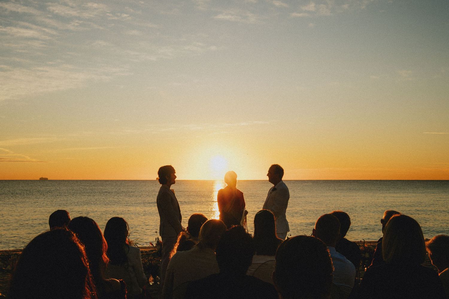 Silhouettes of people gathered on a beach at sunset during an outdoor ceremony with golden ocean backdrop.