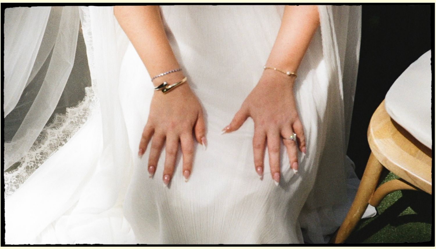 Bride's hands resting on white wedding dress, wearing gold bracelets and a ring, with soft natural light.