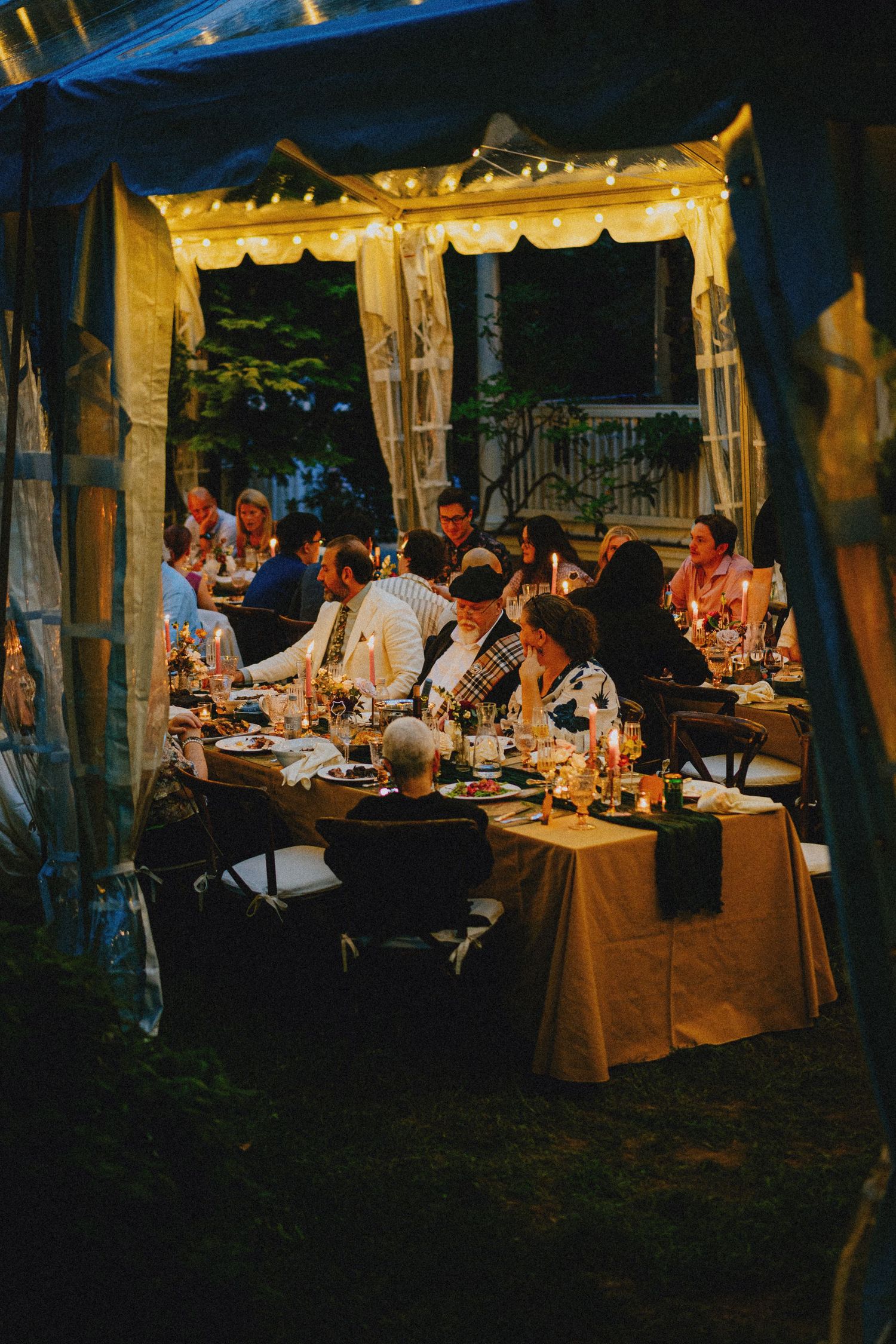 Guests dining under a warmly lit outdoor tent with string lights and candles at an elegant evening event.