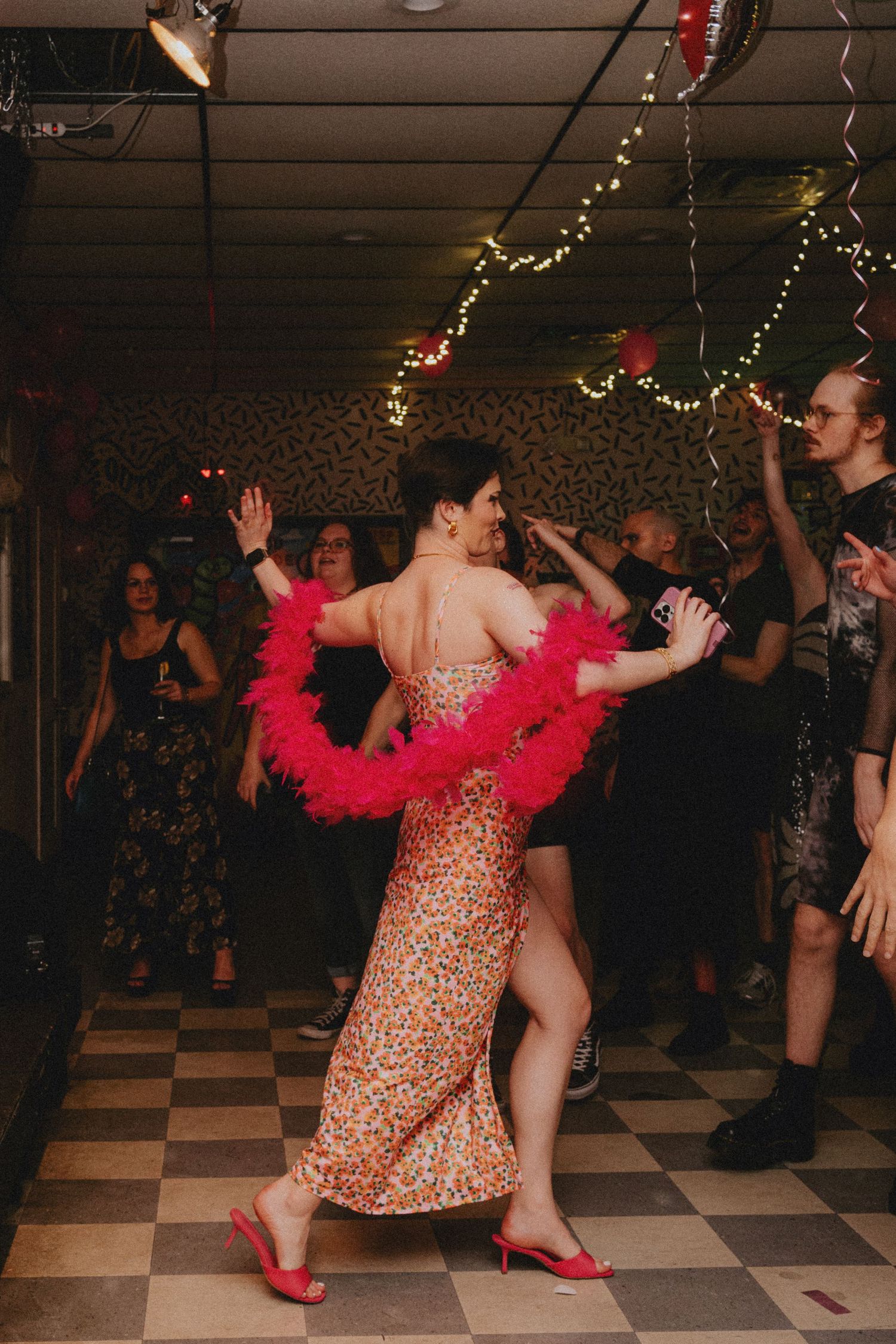 Woman in floral dress and pink feather boa dancing at a party with string lights and checkered floor.