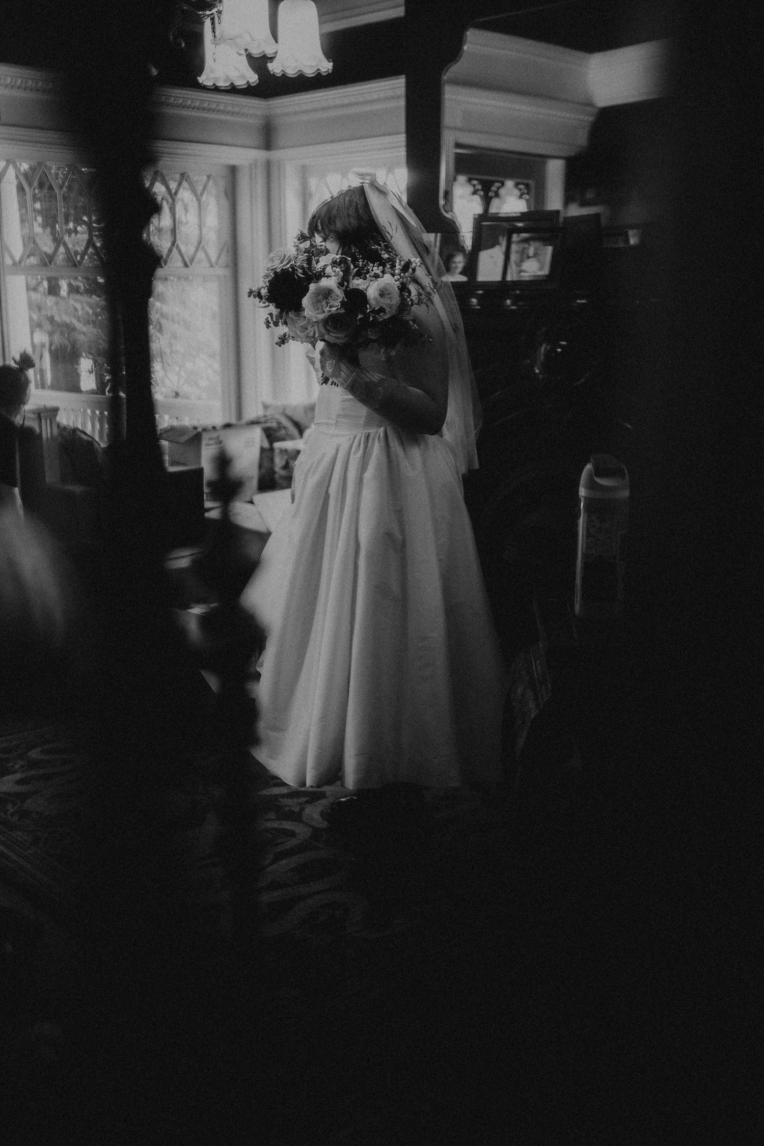 Black and white photo of a bride in a white gown holding a bouquet, standing in an elegant dimly lit room.