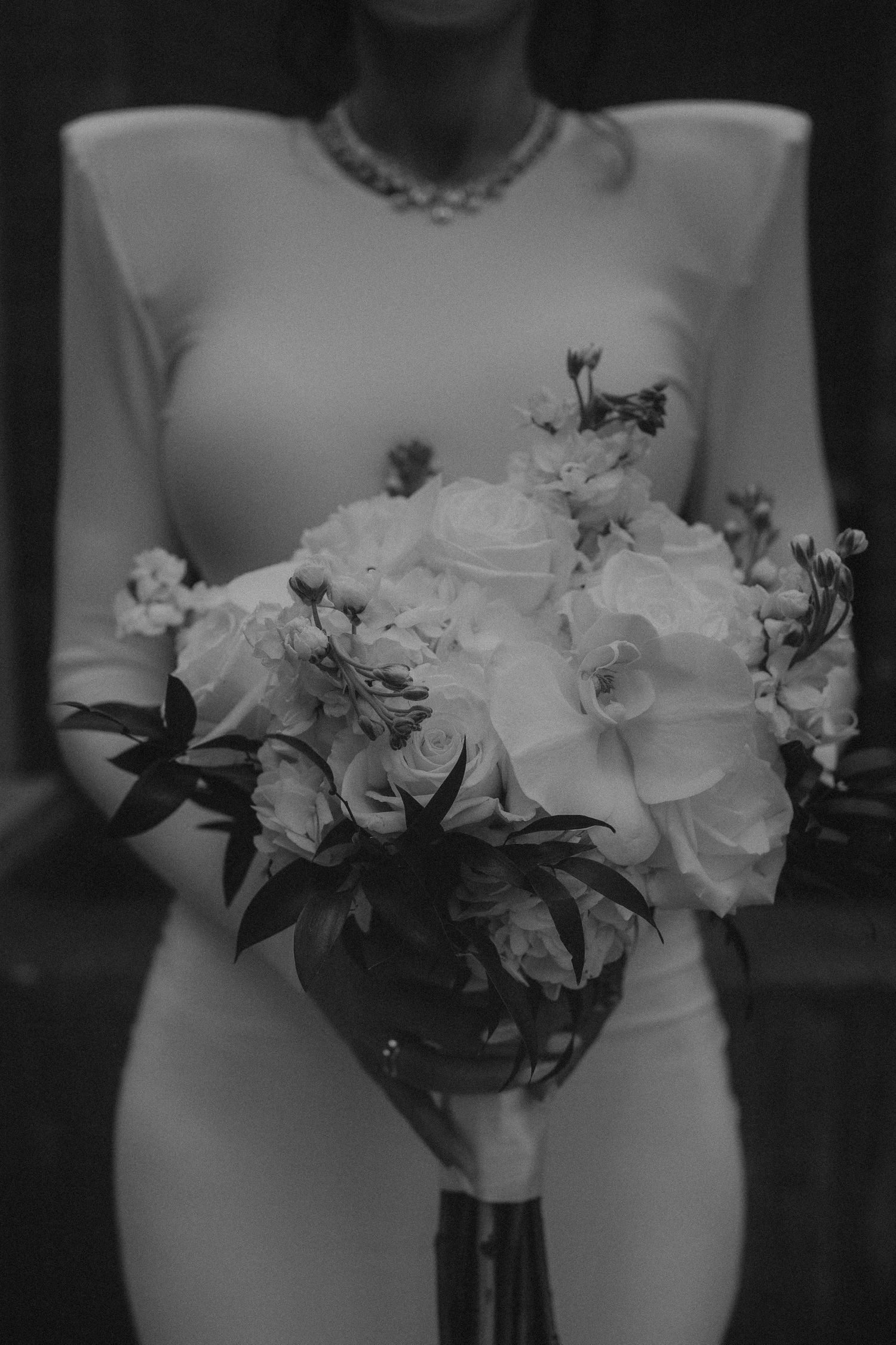 Black and white photo of a bride in a fitted gown holding a lush bouquet of roses and orchids.