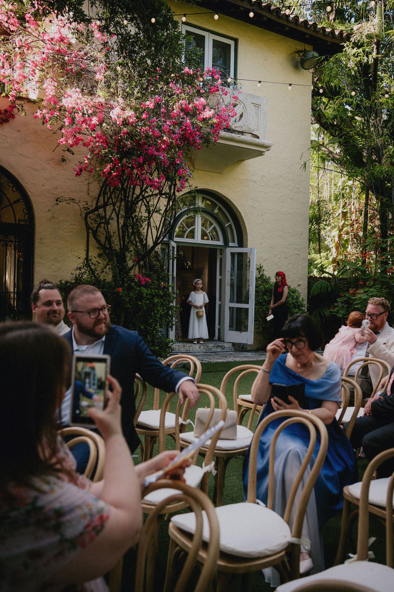 Guests seated on wooden chairs in a garden courtyard during an outdoor wedding ceremony at a yellow villa.