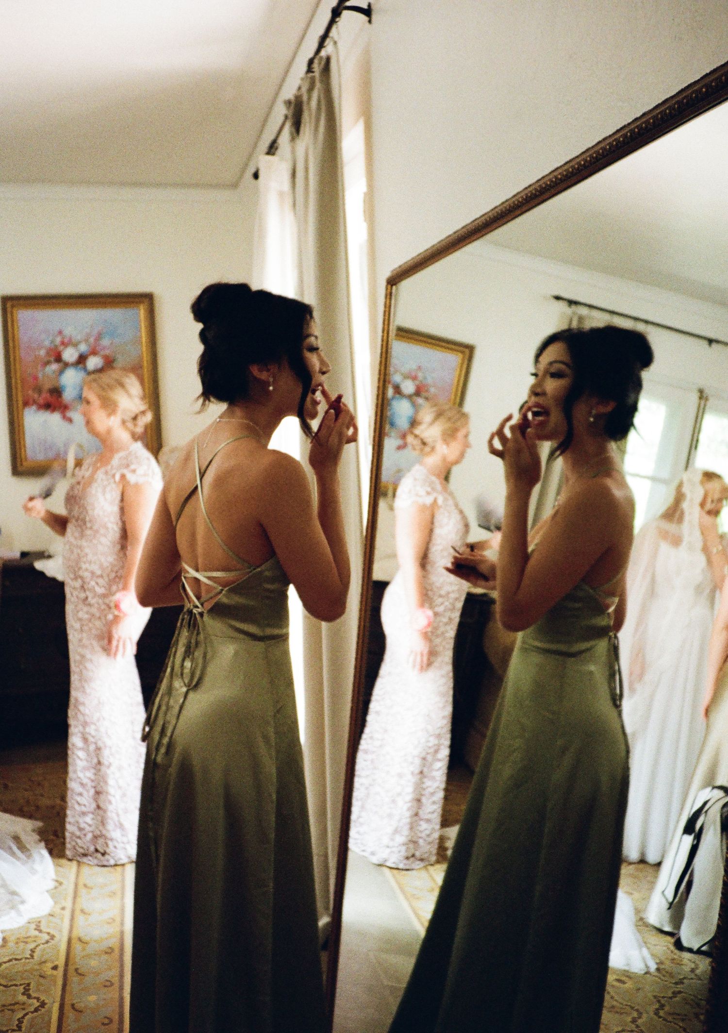 Bridesmaid in olive green gown applying makeup before mirror at wedding preparation.