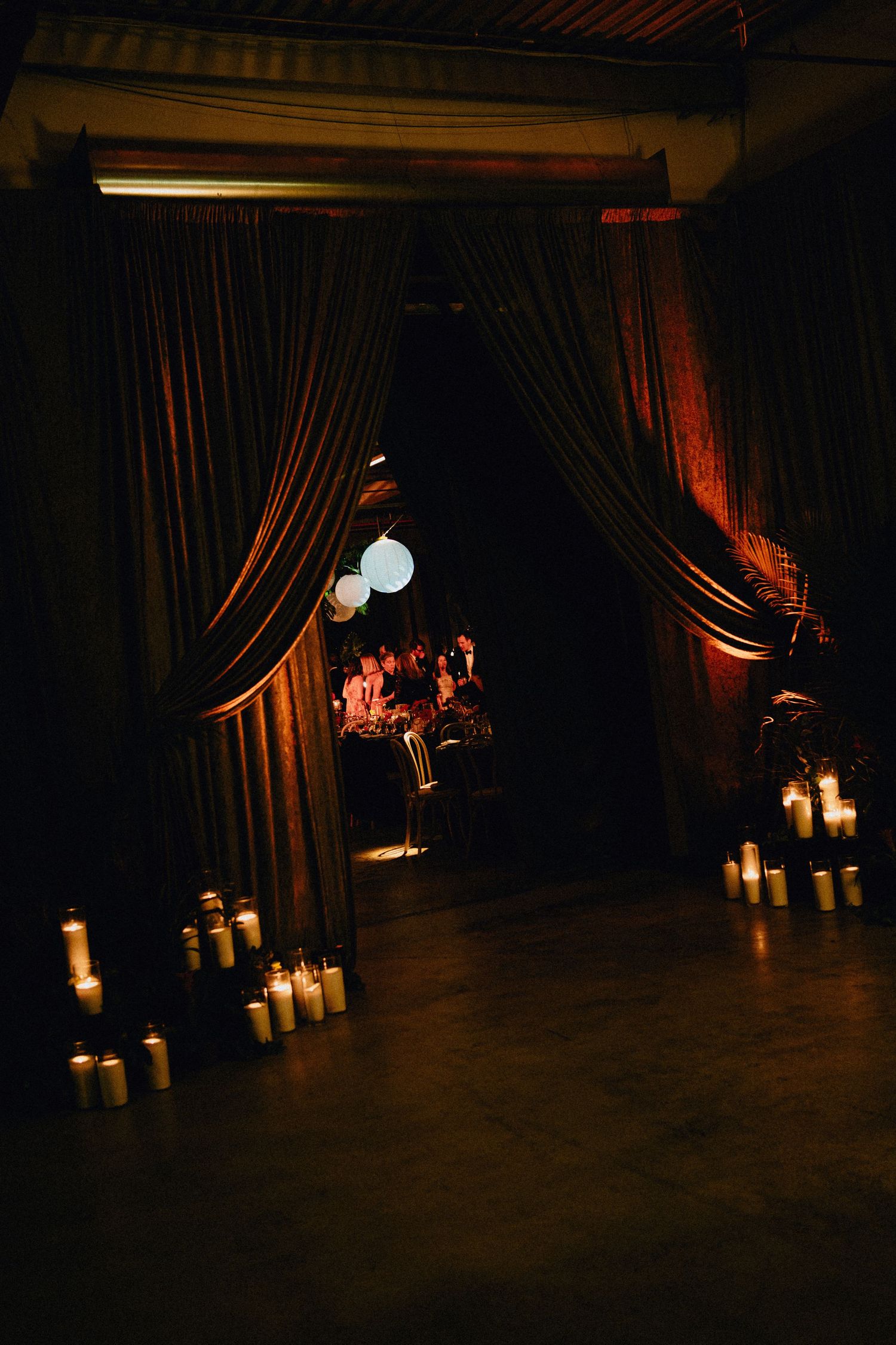 Dramatic dark event space with draped curtains, glowing candles lining the floor, and paper lanterns illuminating an intimate dinner beyond.