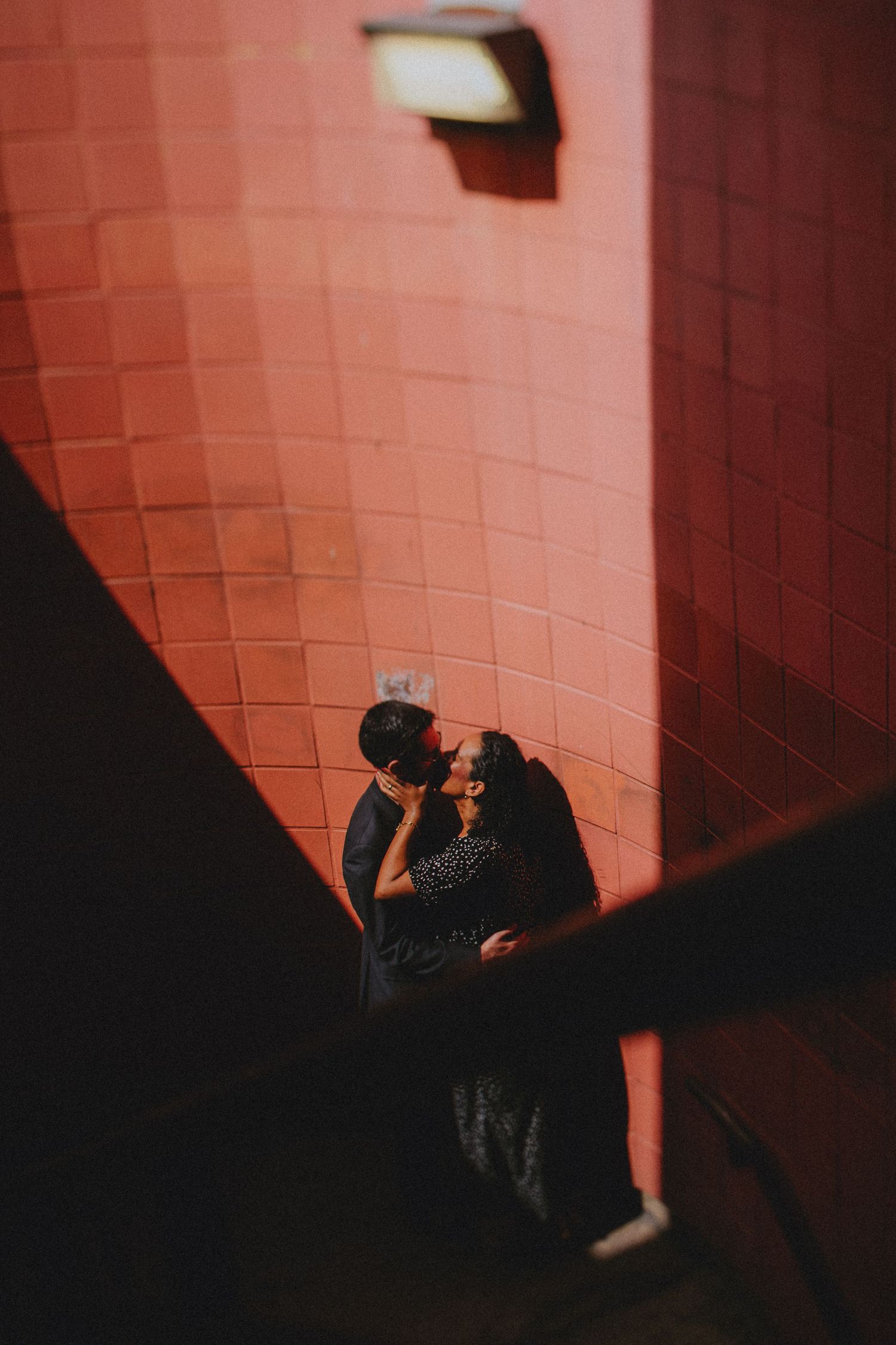 Couple sharing a romantic kiss against a red tiled wall, viewed from above in dramatic shadow and light.
