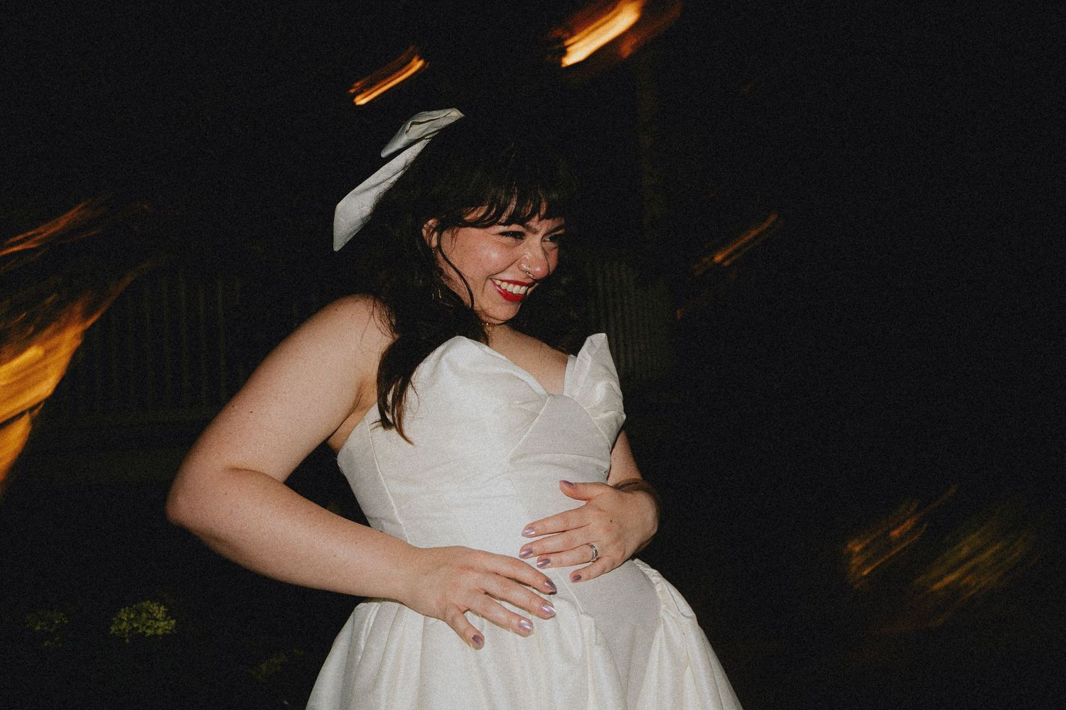 Bride in white wedding dress and bow hair accessory smiling and dancing joyfully at an outdoor evening reception.
