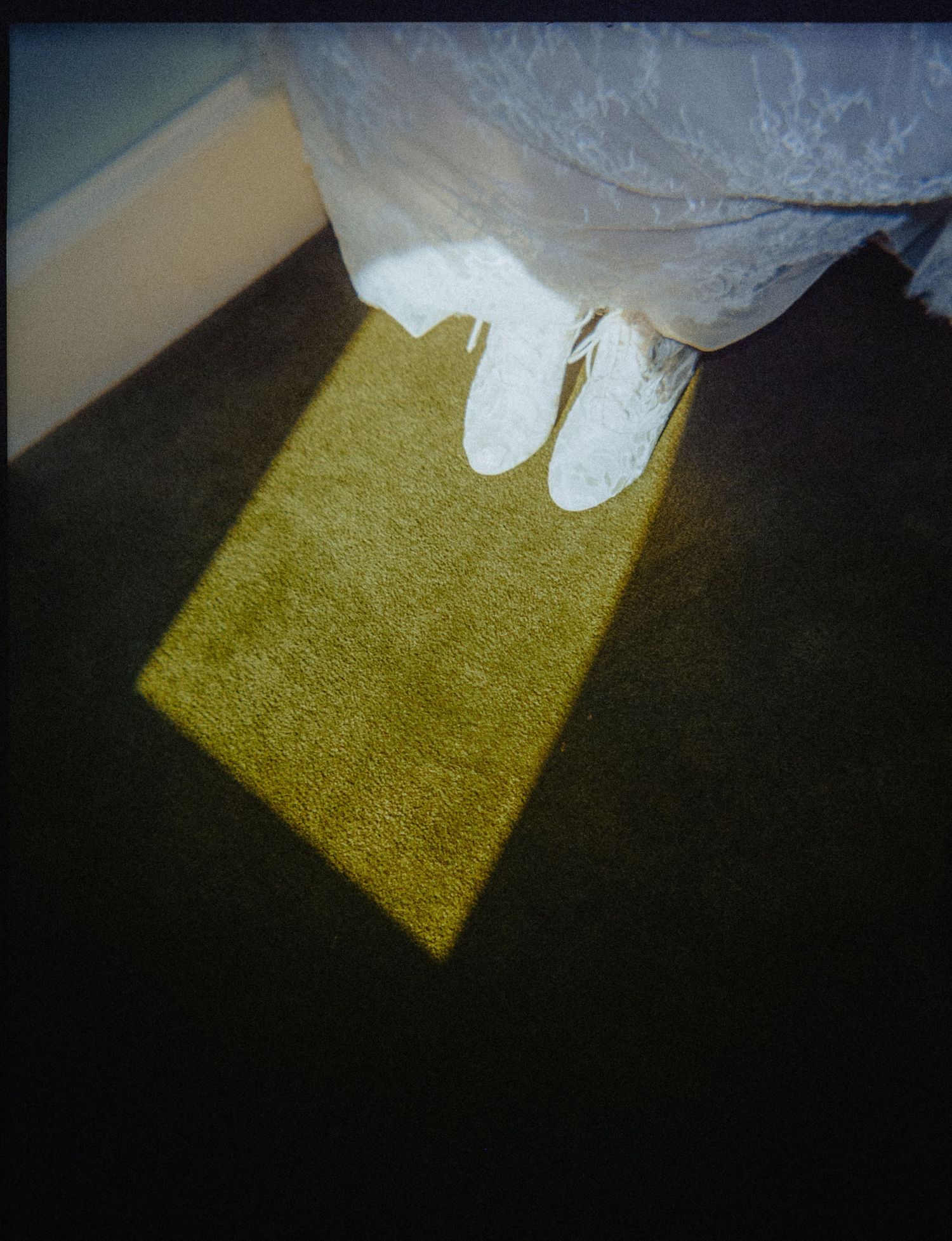 A bride in white lace dress and white shoes stands on a sunlit yellow-green carpet patch against dark flooring.