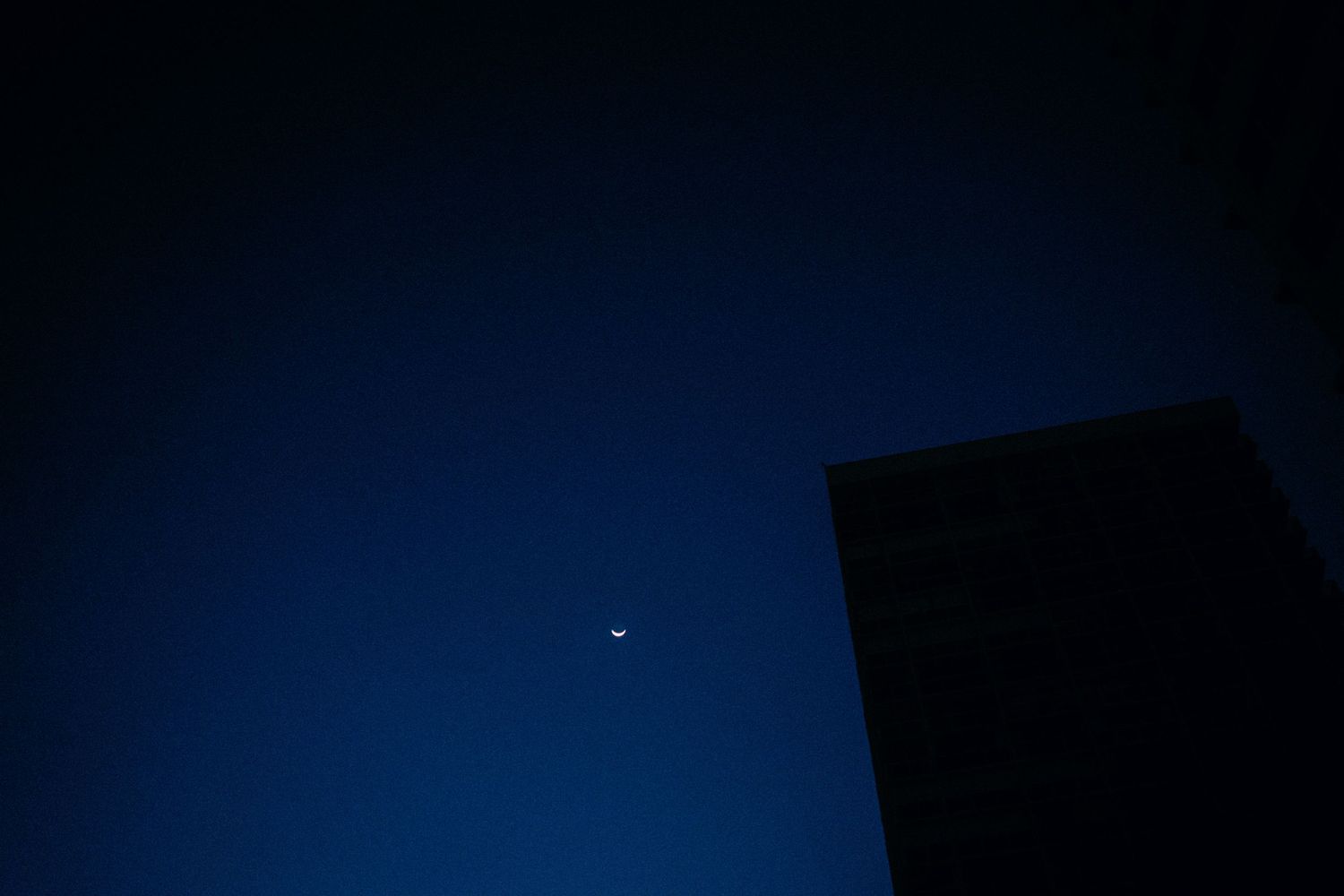 A crescent moon glows in a deep blue twilight sky above a dark silhouetted building corner.