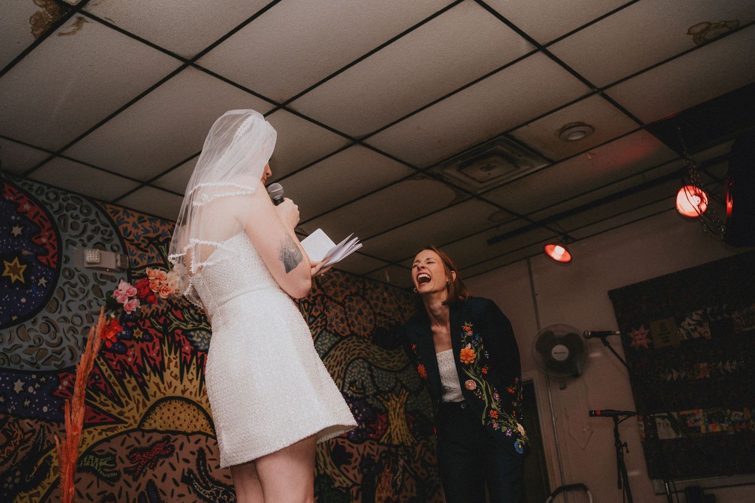 Bride in white mini dress and veil reads vows while officiant laughs during indoor wedding ceremony with colorful tapestry backdrop.