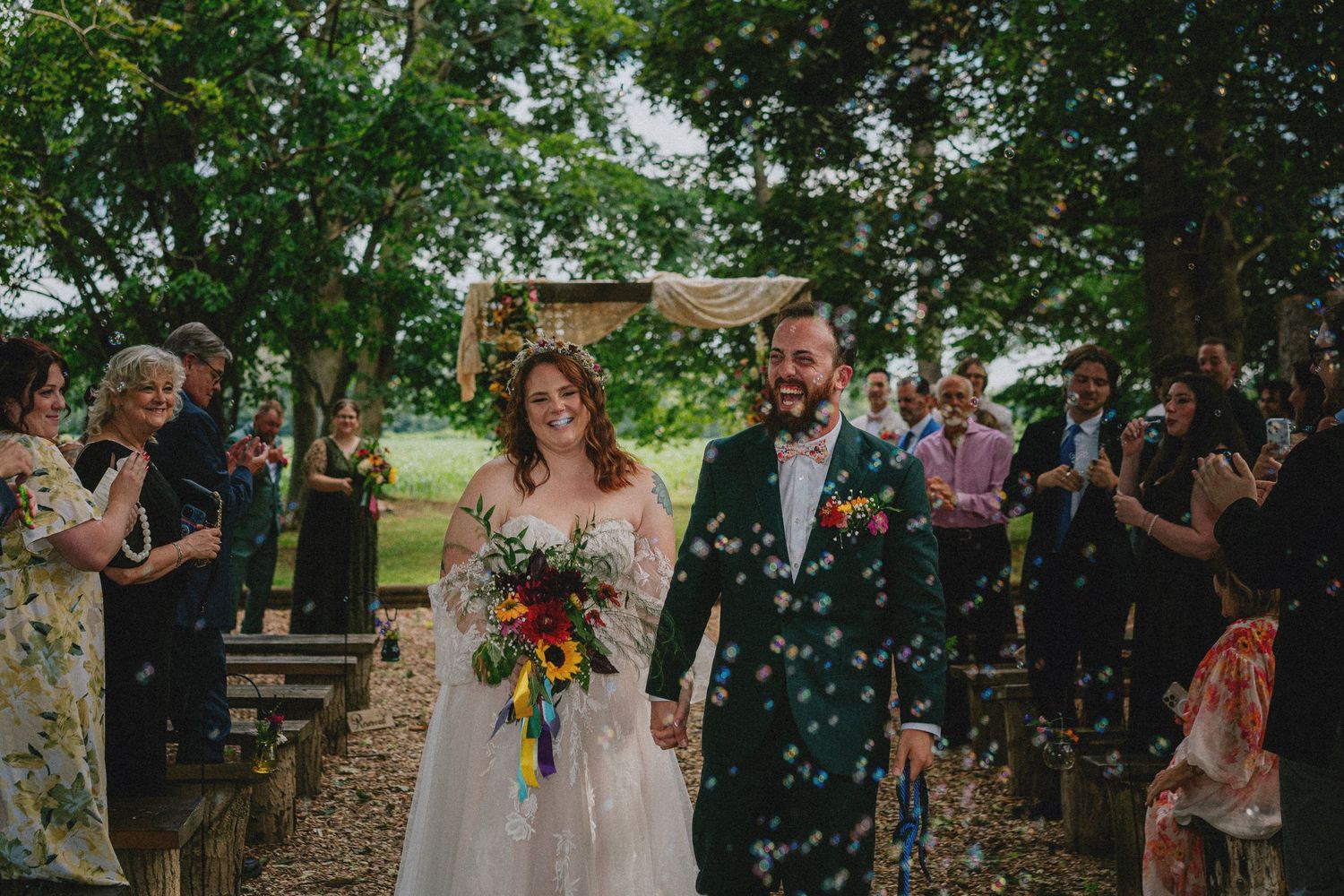Bride and groom walk down outdoor aisle as guests toss bubbles at rustic woodland wedding ceremony.