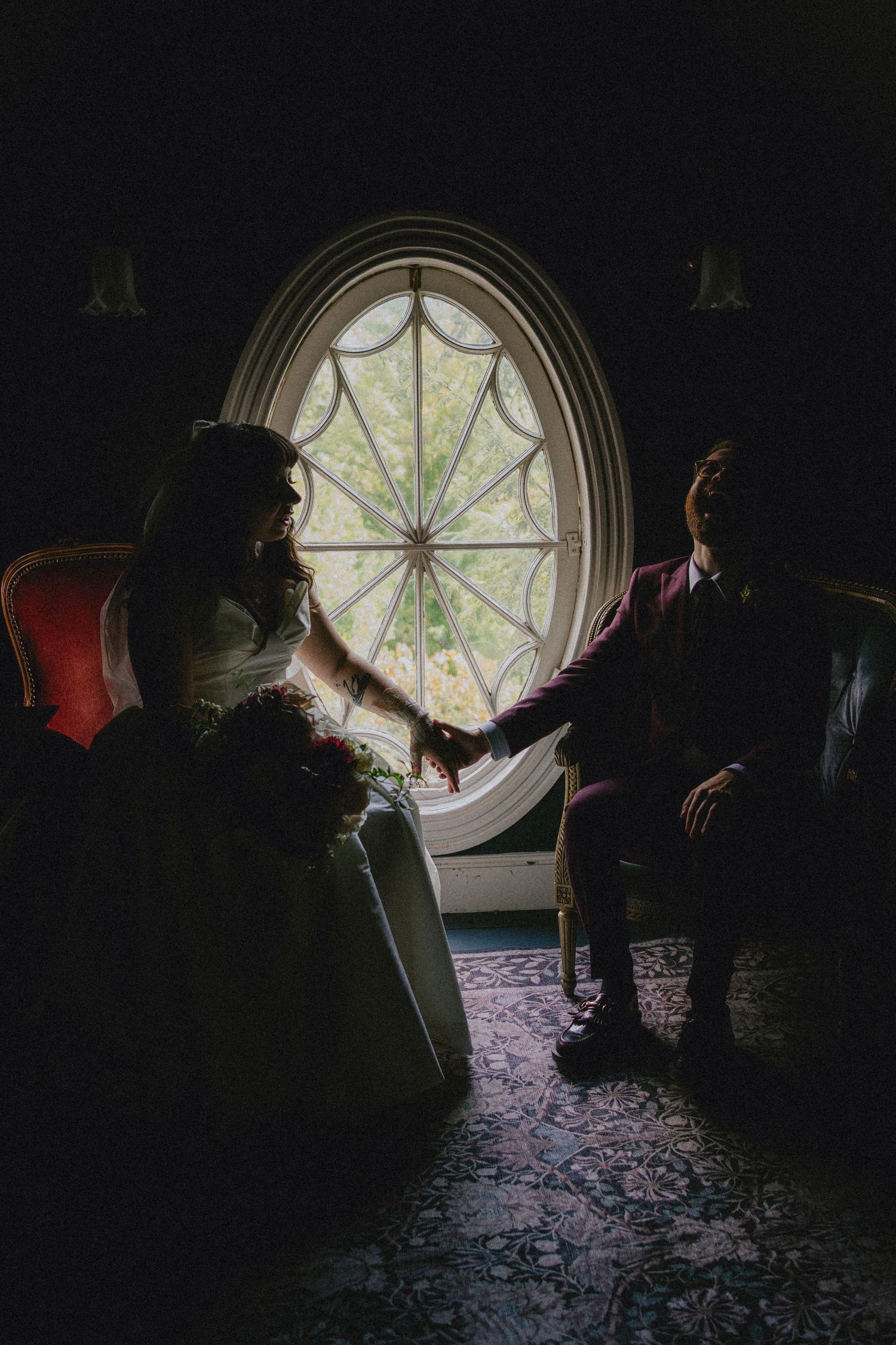 Silhouette of bride and groom holding hands by an oval stained glass window in a dimly lit elegant room.