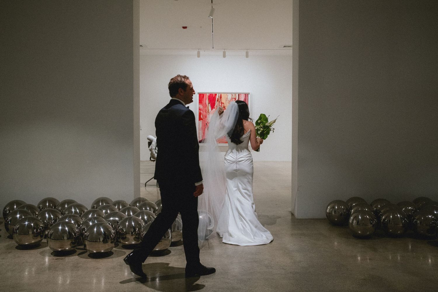Bride and groom walking through elegant wedding venue with silver balloon decor and modern art on white walls.