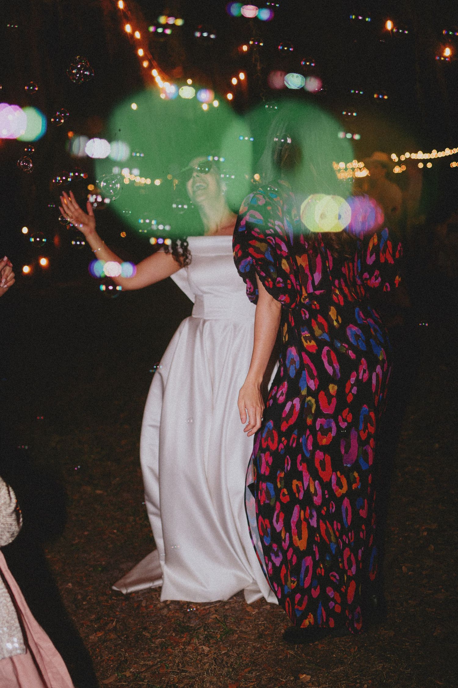 Bride in white gown dancing outdoors at night with guest in colorful leopard print dress, bokeh lights glowing.