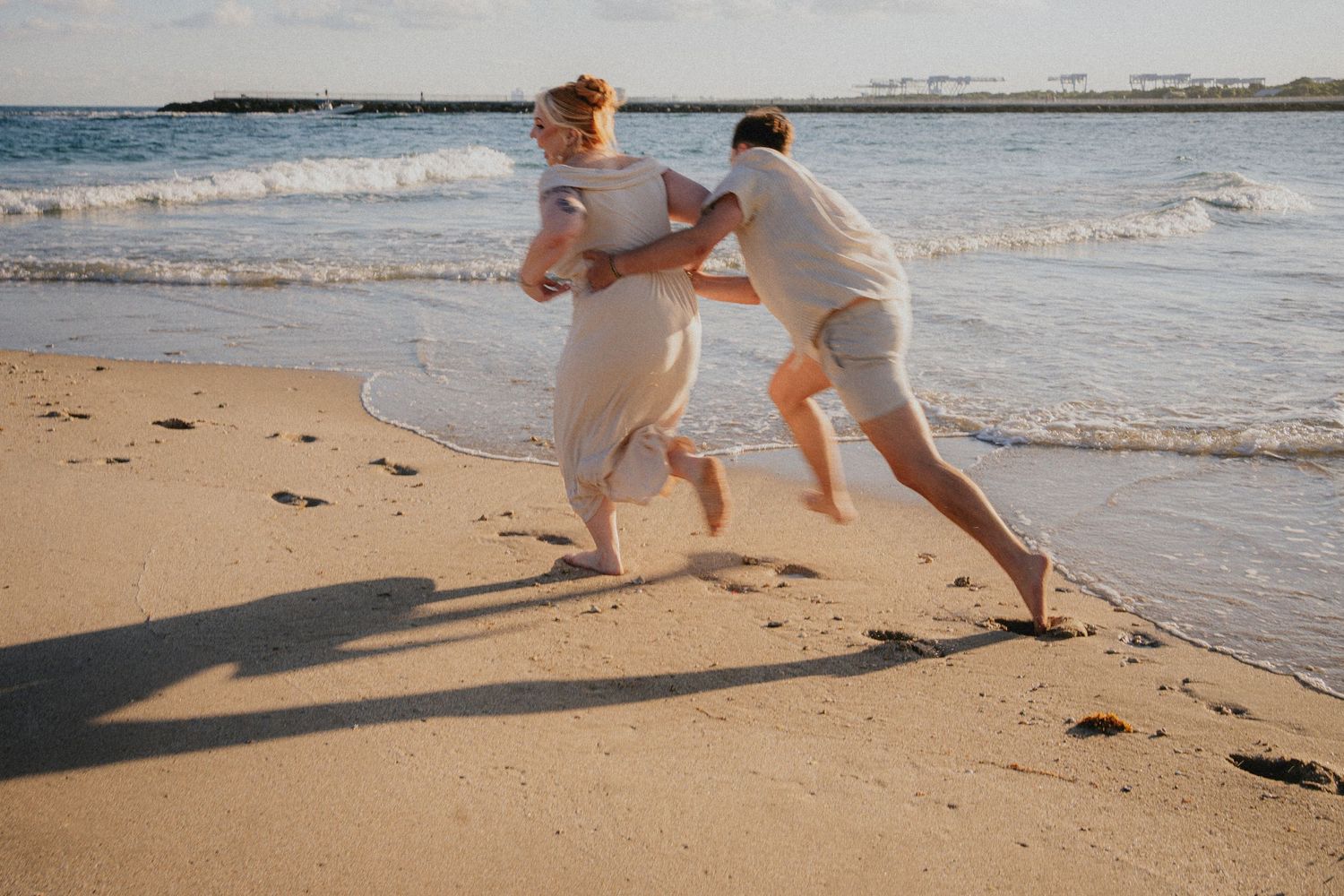 Couple playing and running together on a sandy beach near ocean waves on a sunny day.