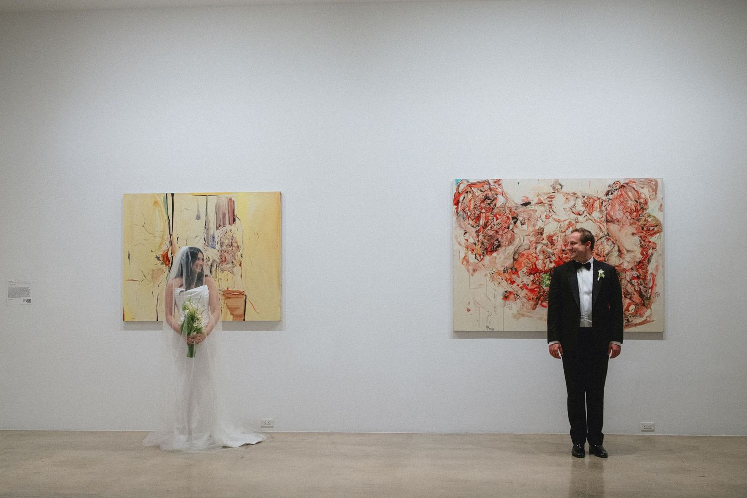 Bride and groom standing apart in modern art gallery with abstract paintings on white walls during wedding celebration.