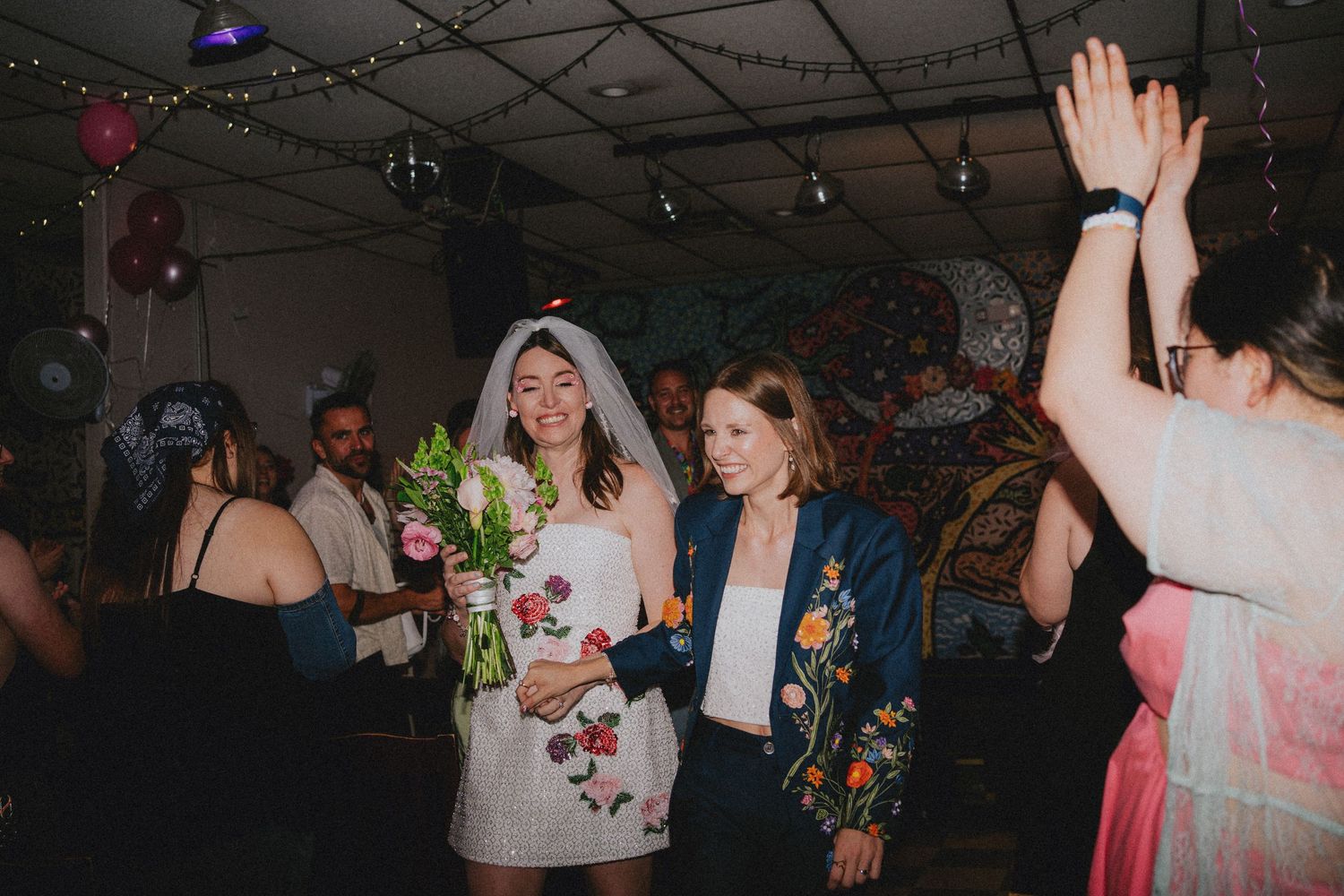 Bride in short white dress with floral bouquet dancing with guests at a colorful wedding reception party.