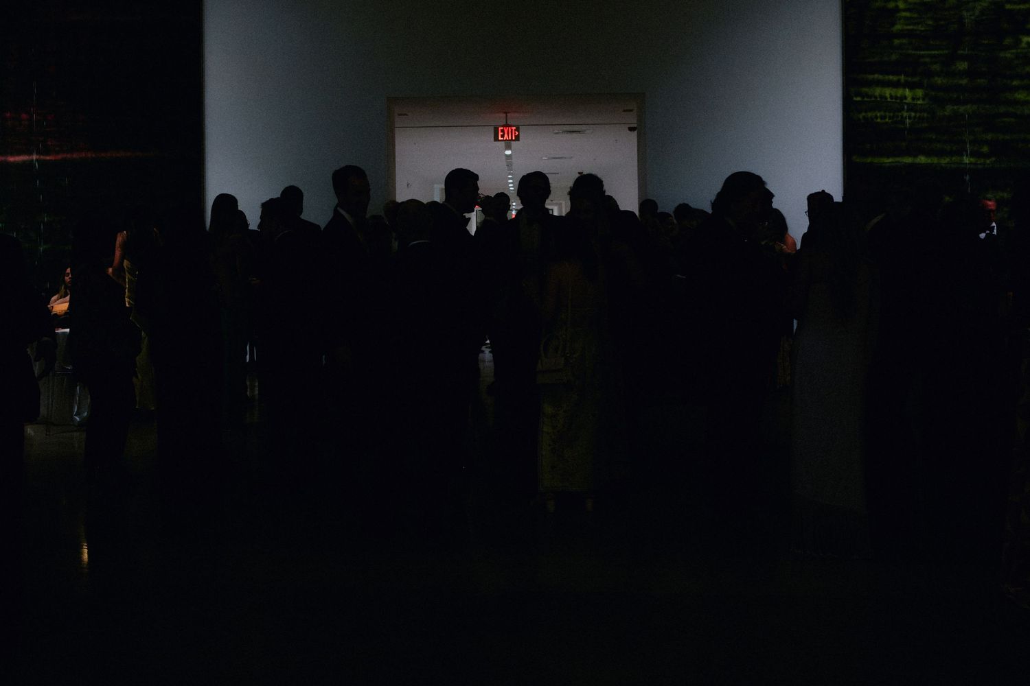 Silhouettes of a crowd gathered in a dark room facing a bright illuminated doorway with a red exit sign.