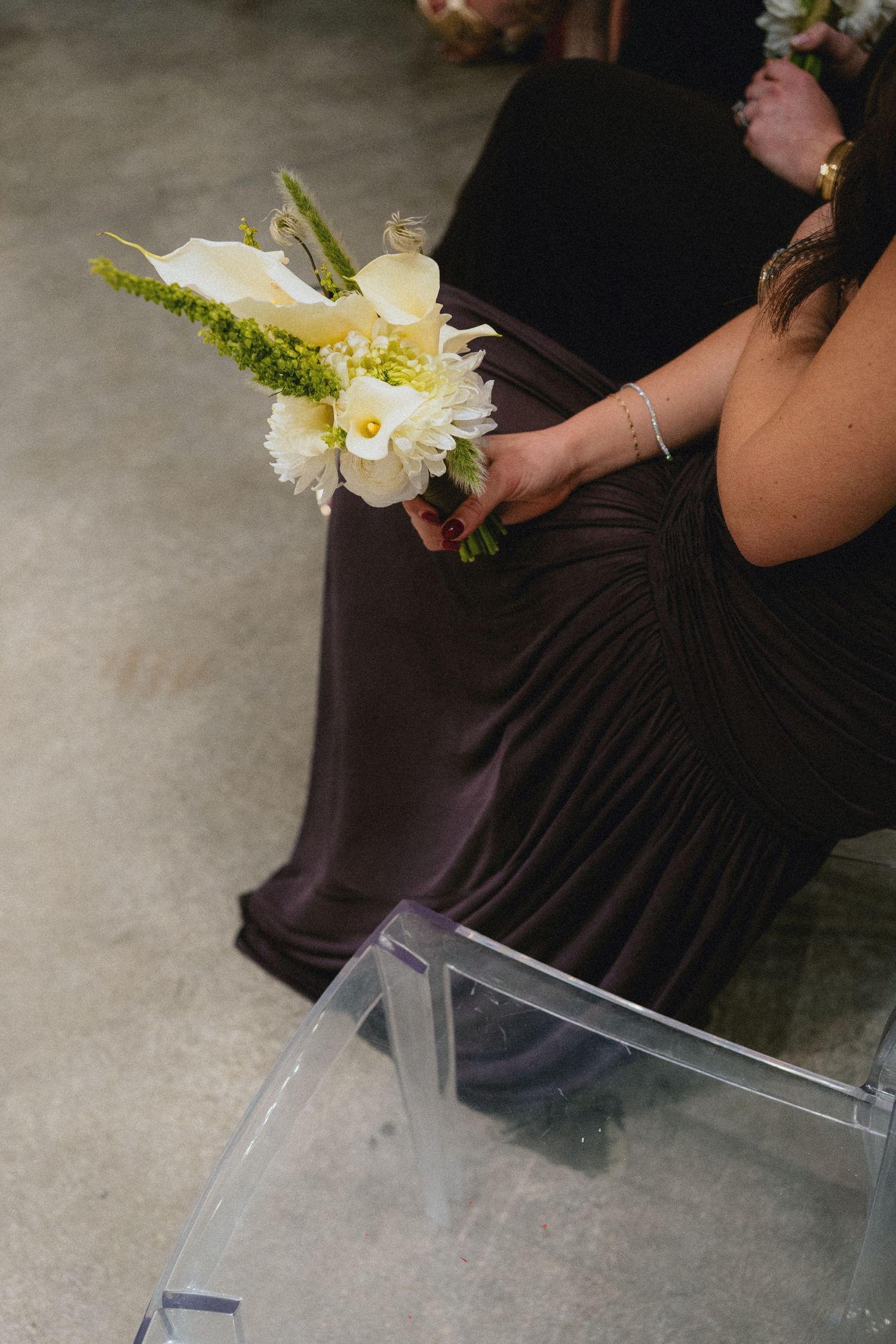 Bridesmaid in dark purple gown holding white floral bouquet with yellow accents while seated on clear acrylic chair.