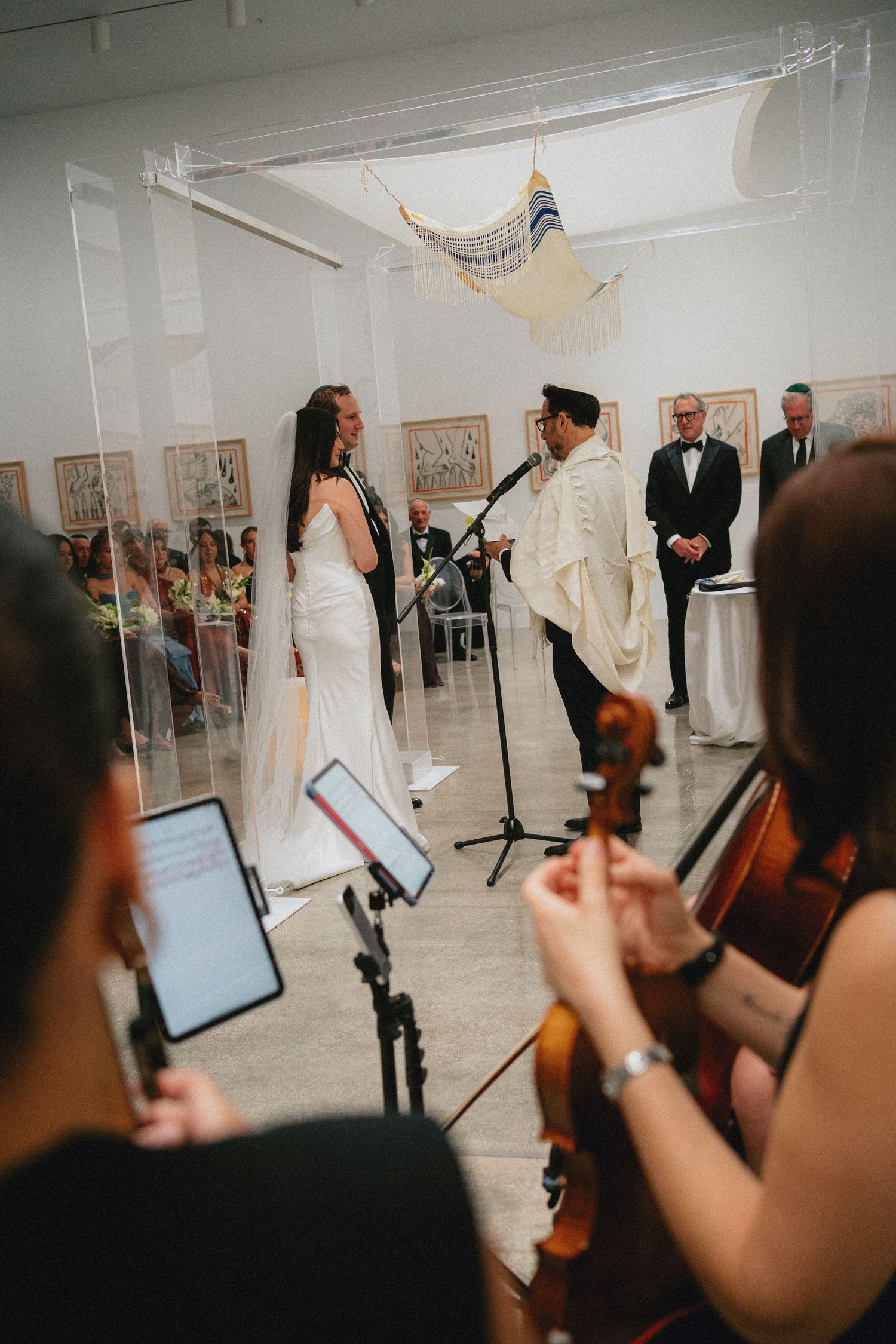 Jewish wedding ceremony in art gallery with bride, groom, and officiant under chuppah, with string musicians performing in foreground.