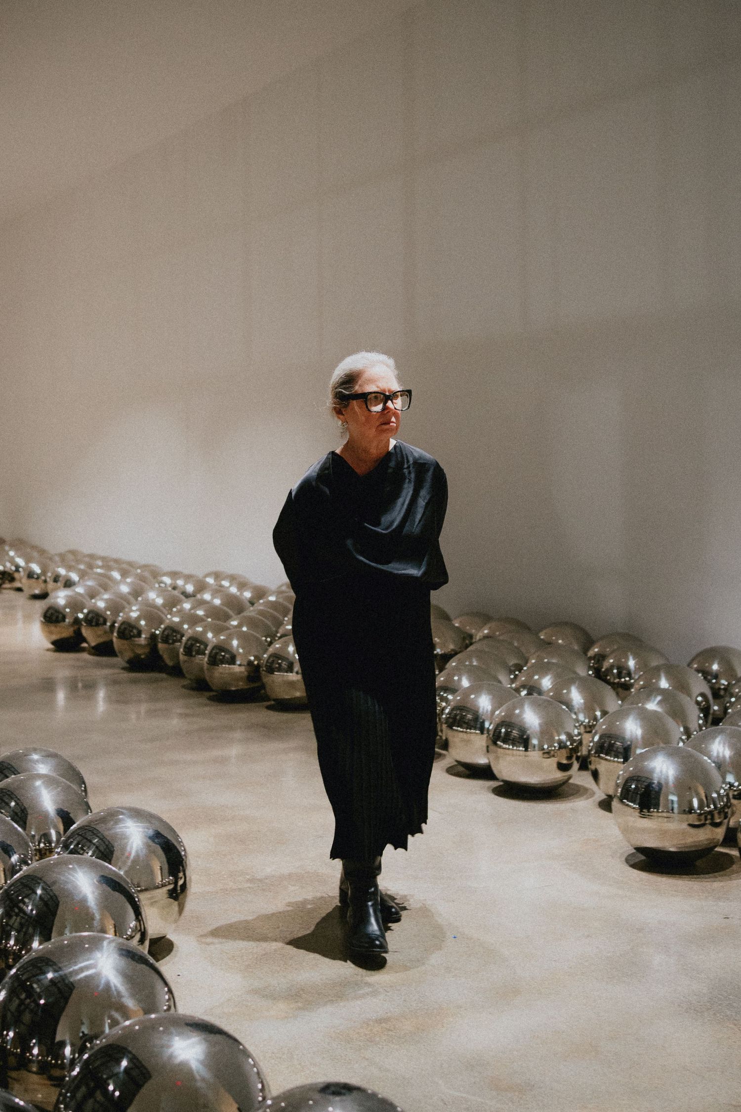 Woman in black outfit stands among shiny silver mirror balls in a modern gallery space.