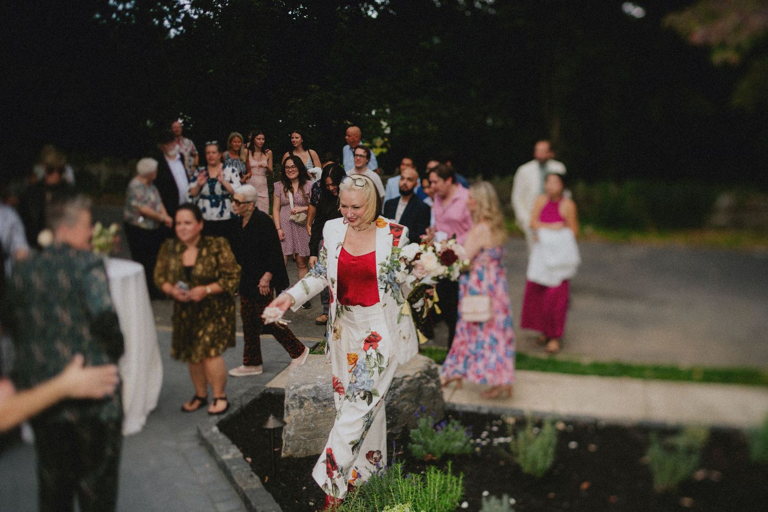 Bride in floral dress walks past garden pond at outdoor wedding reception with guests gathered in background at dusk.