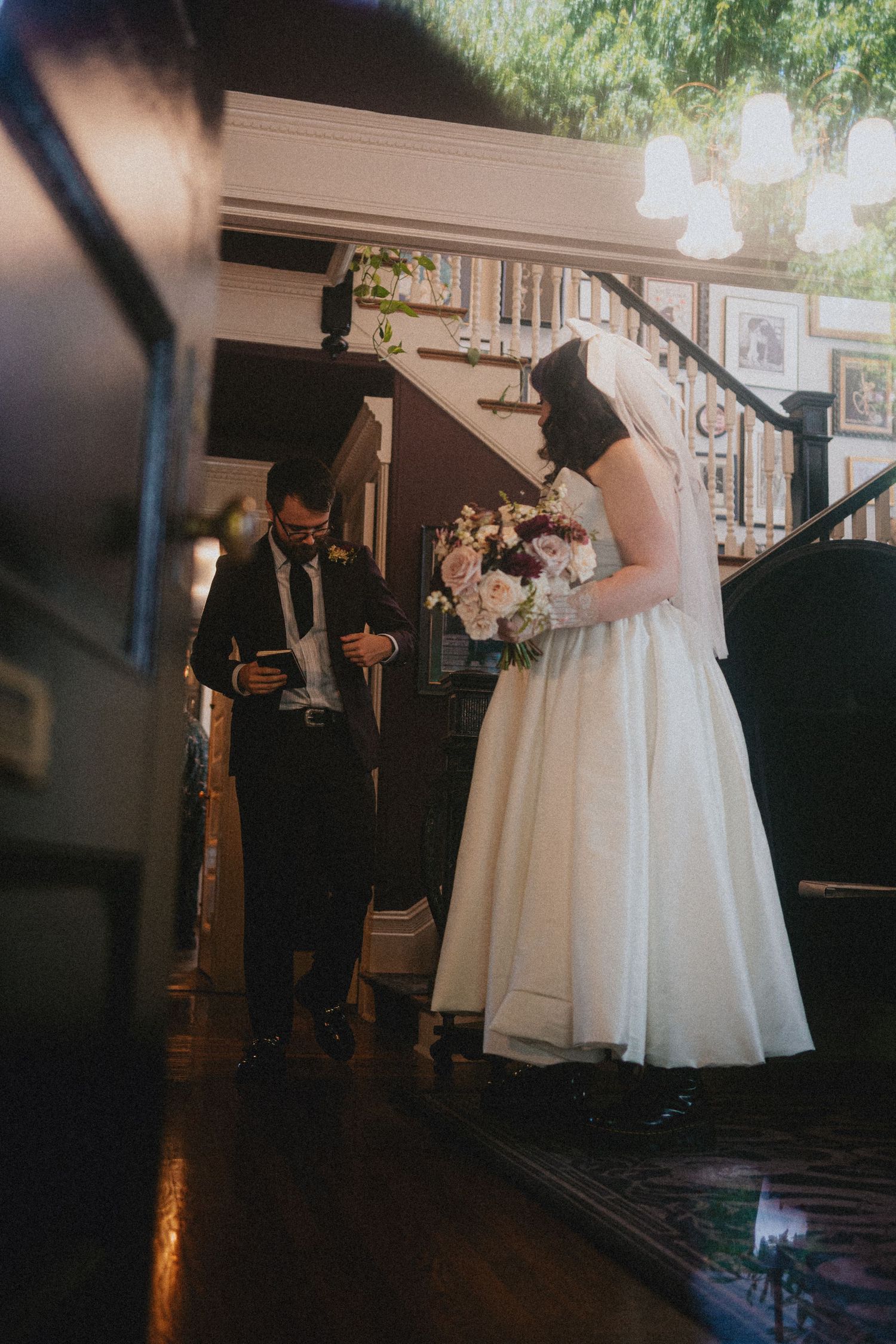 Bride and groom exchanging vows indoors, bride holding pink bouquet, groom reading from card near elegant staircase.
