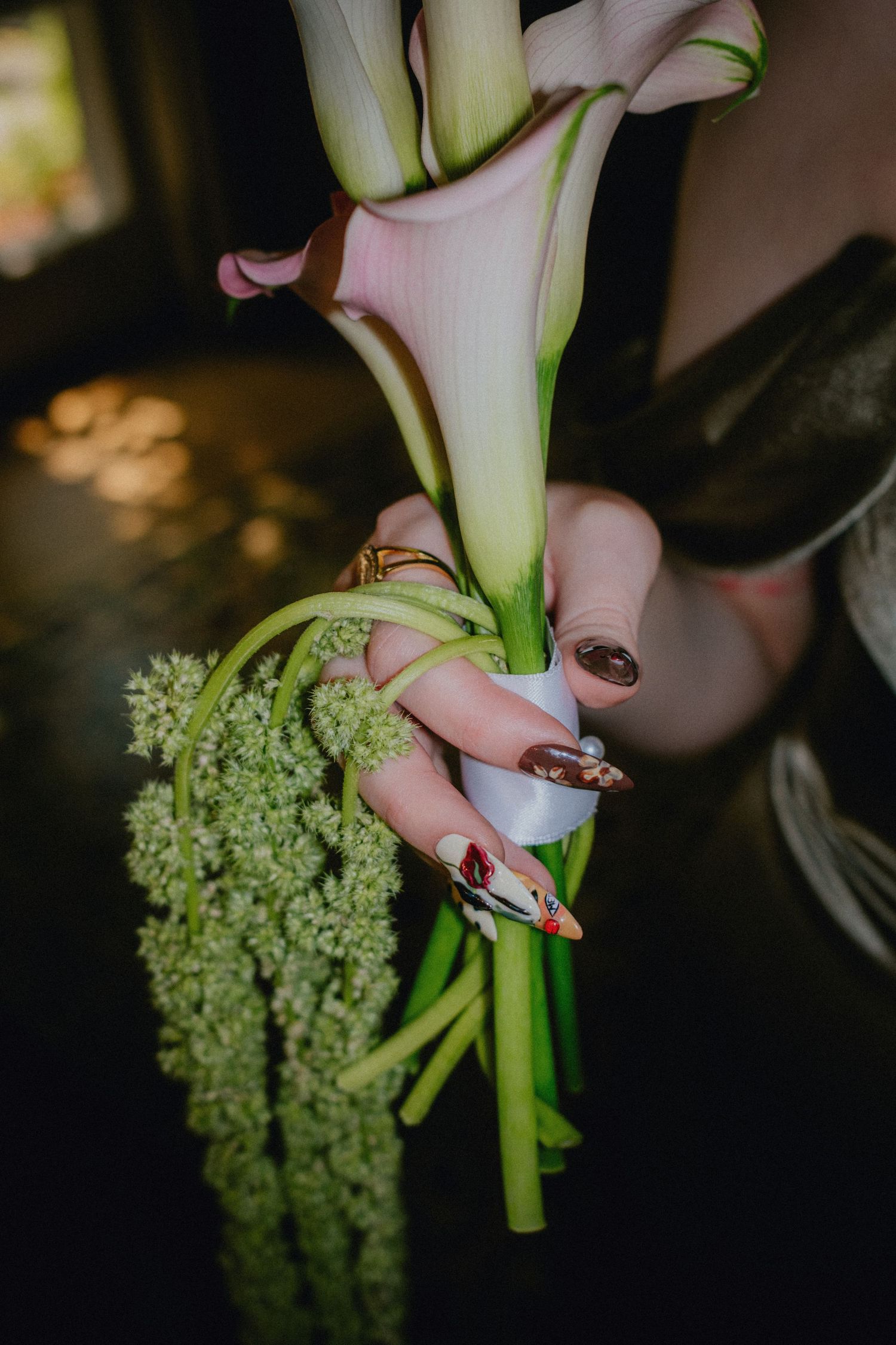 Close-up of hands with long decorative nails holding pink calla lilies and green flowers, adorned with gold rings.
