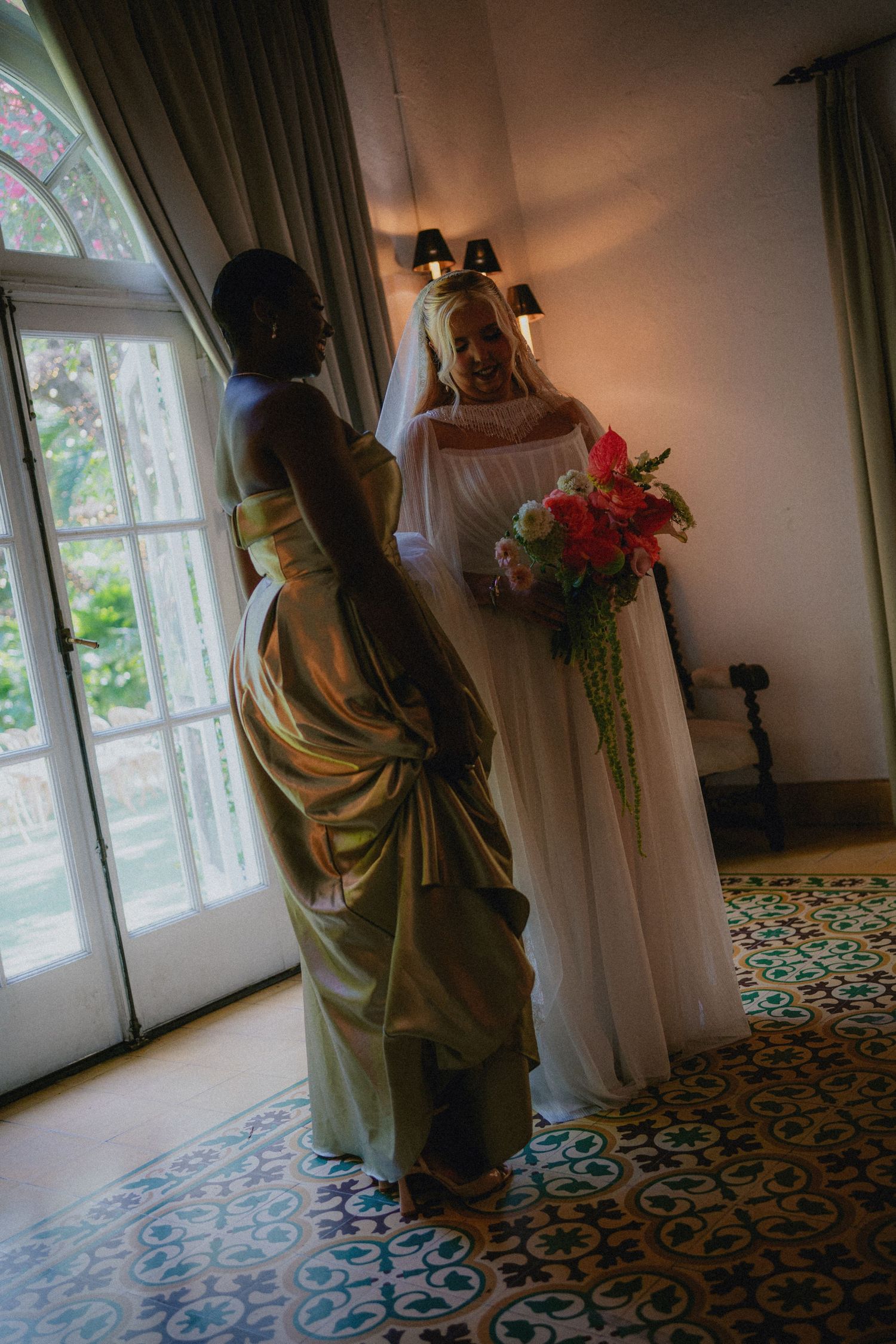 Bride in white gown holding colorful bouquet stands with bridesmaid in gold dress near French doors.