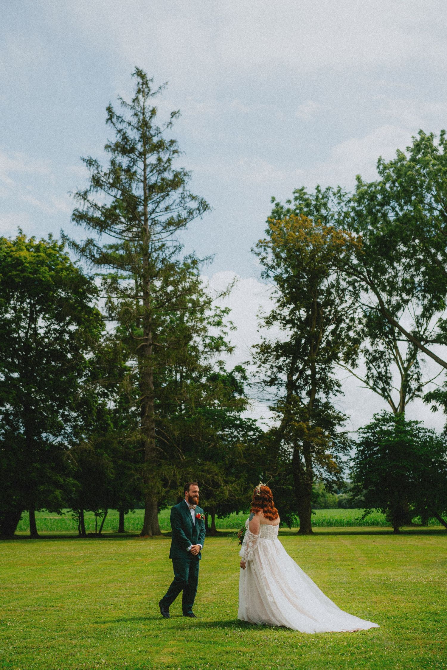 A bride and groom share a romantic moment on a lush green lawn surrounded by tall trees on their wedding day.